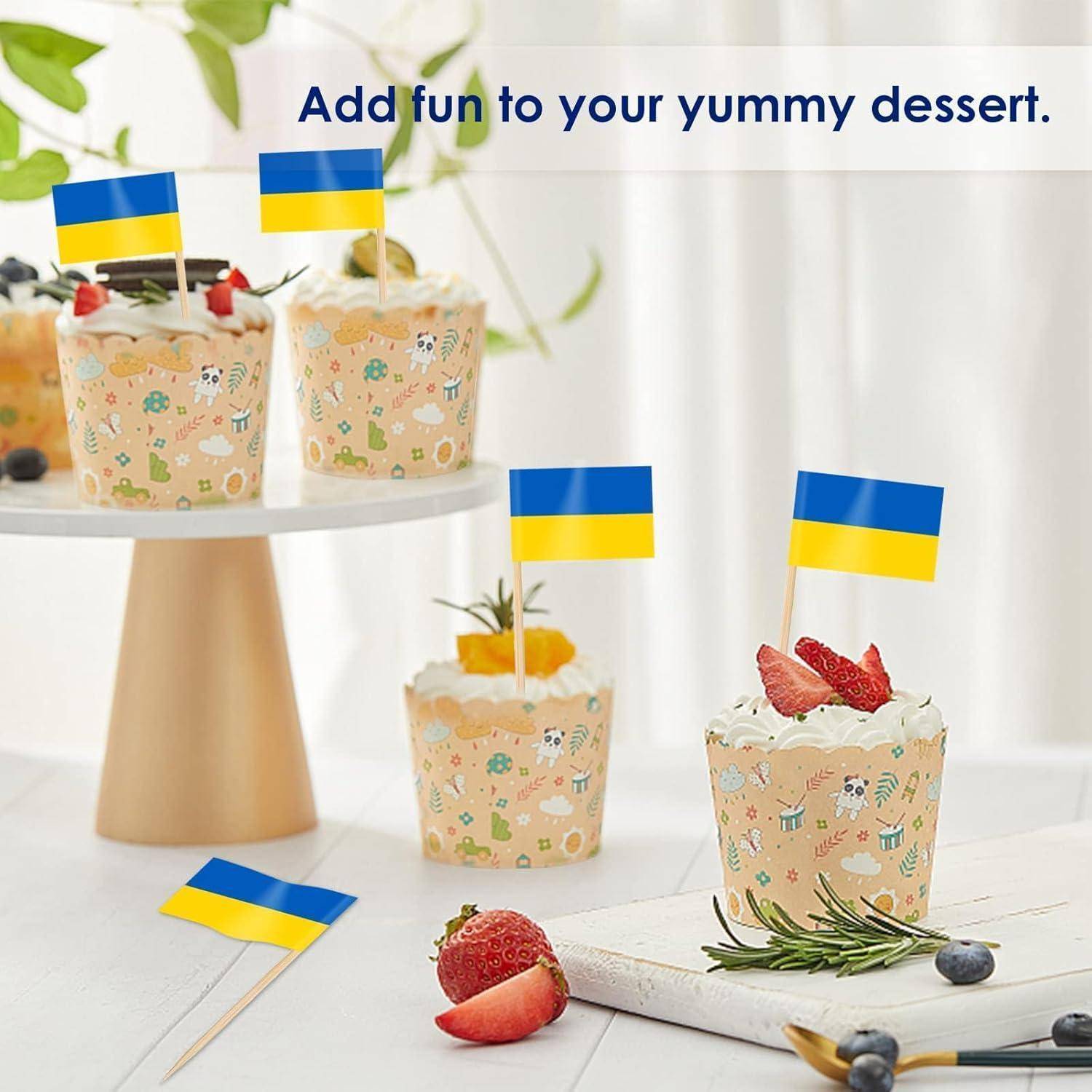 Drei Cupcakes, die mit Zahnstocherfähnchen in Blau und Gelb – den Farben der ukrainischen Flagge – gekrönt sind. Eine Botschaft lautet: „Gib deinem leckeren Dessert mehr Spaß.