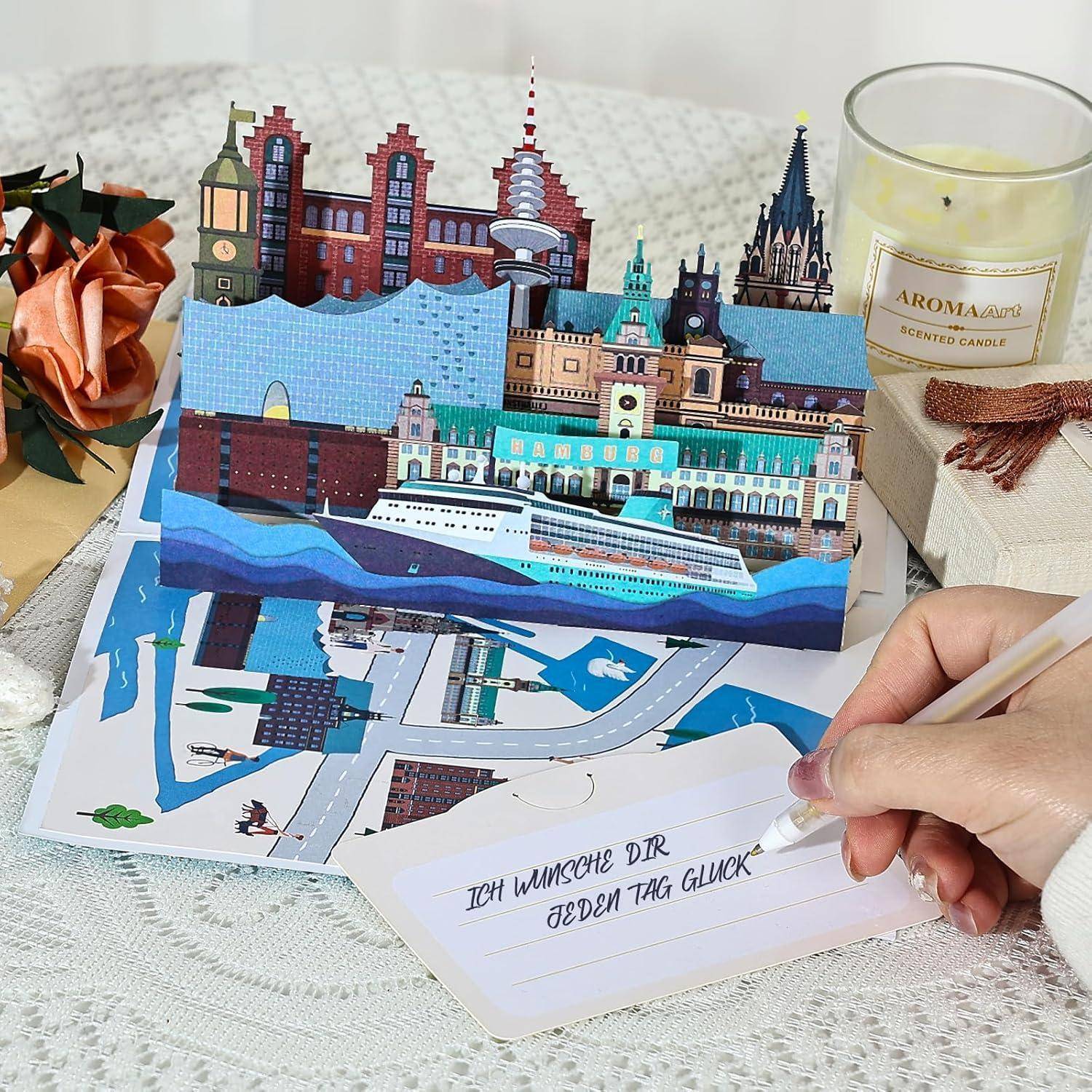 Eine Pop-up-Karte mit europäischen Sehenswürdigkeiten und einem Kreuzfahrtschiff. Handschriftlich ist auf einer Notiz auf Deutsch zu lesen: „Ich wünsche dir jeden Tag Glück