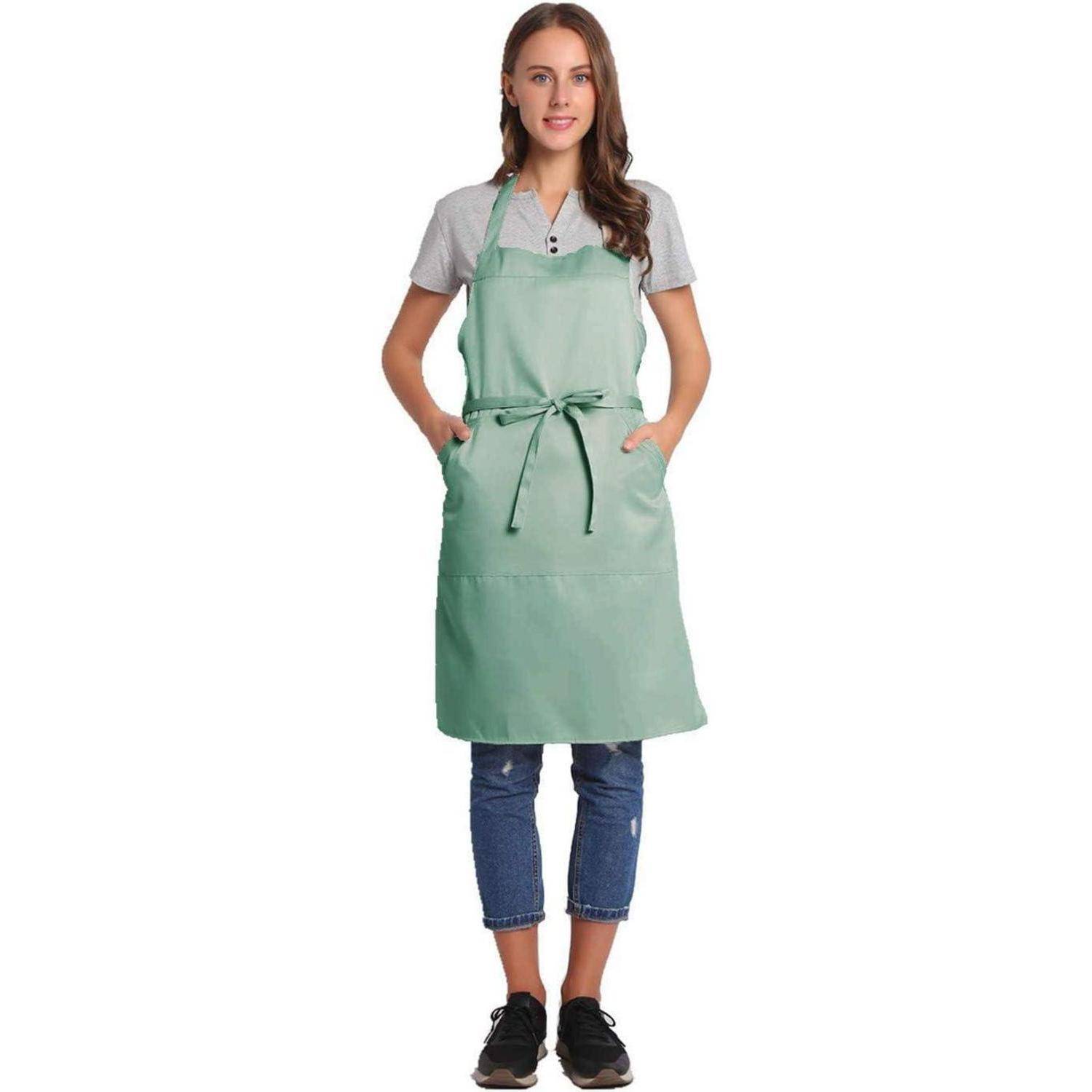 Eine Person trägt eine grüne Schürze, ein graues Hemd und Jeans und steht frontal, die Hände in den Taschender Schürze, vor einem einfarbigen weißen Hintergrund.