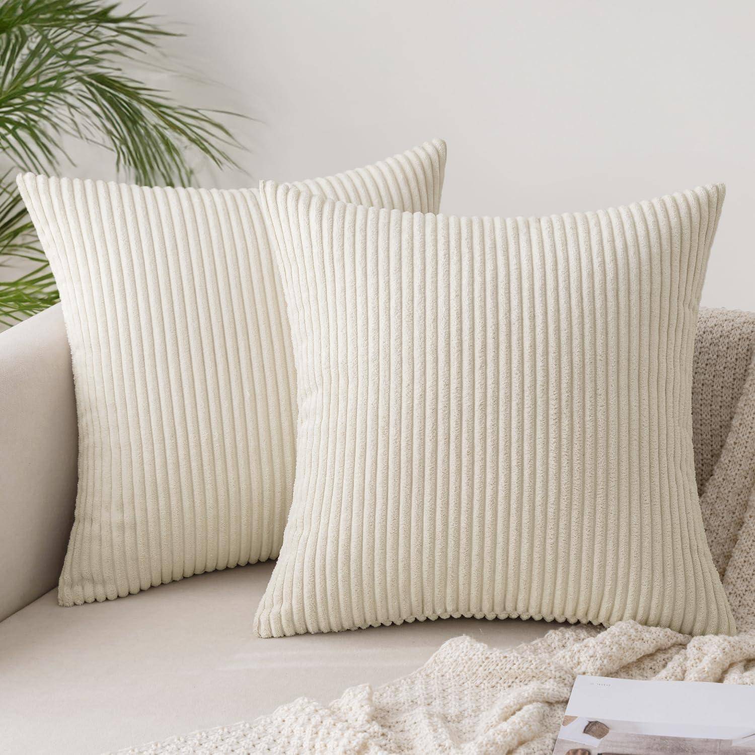 Zwei beige, gerippte Zierkissen auf einem hellgefärbten Sofa mit einer gestrickten Decke, neben grünen Pflanzenblättern.