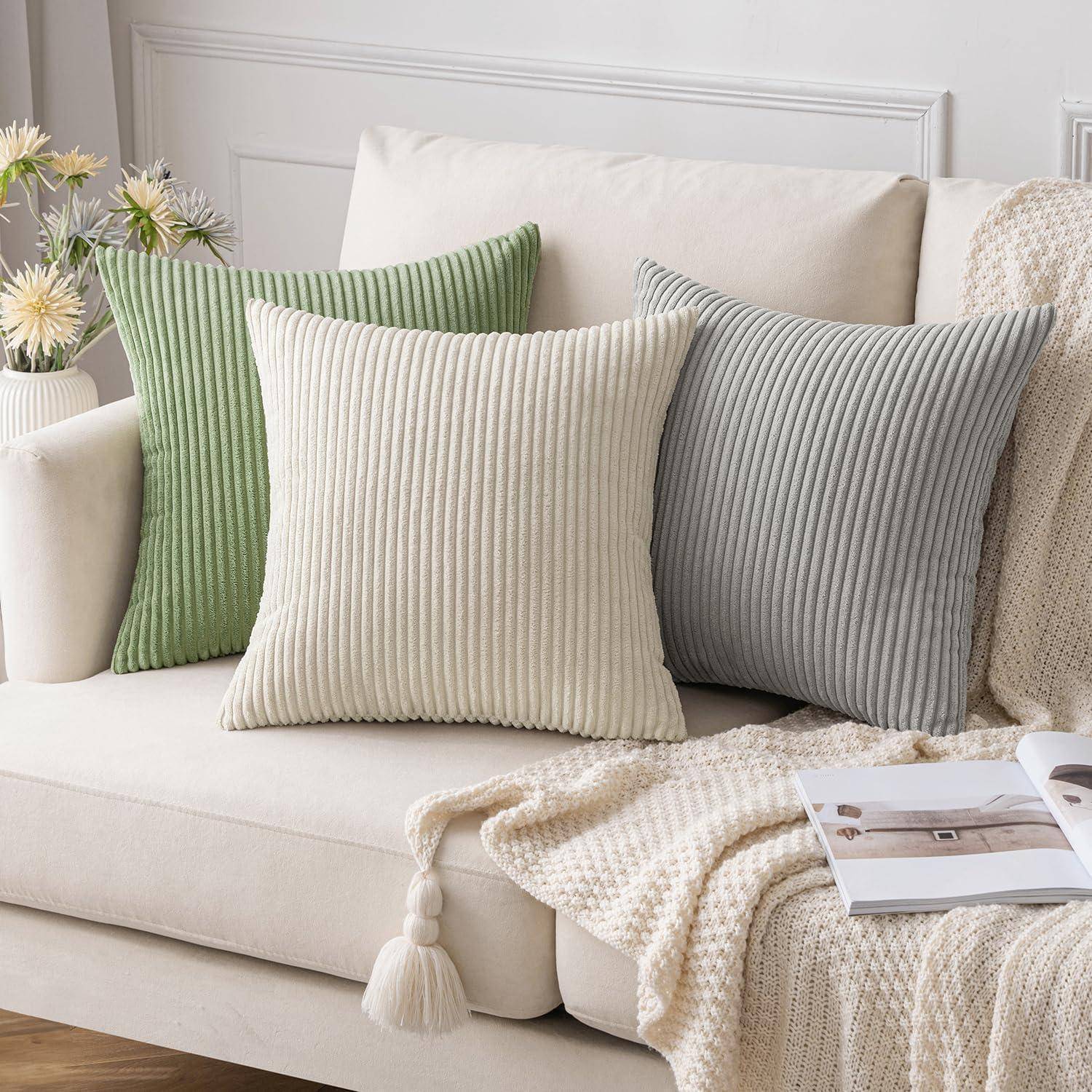 Ein beige Sofa mit grünen, cremefarbenen und grauen gerippten Kissen. Eine gewebte Decke mit Quasten ist über die Armlehne drapiert. Eine Vase mit Blumen schmückt die Seite.