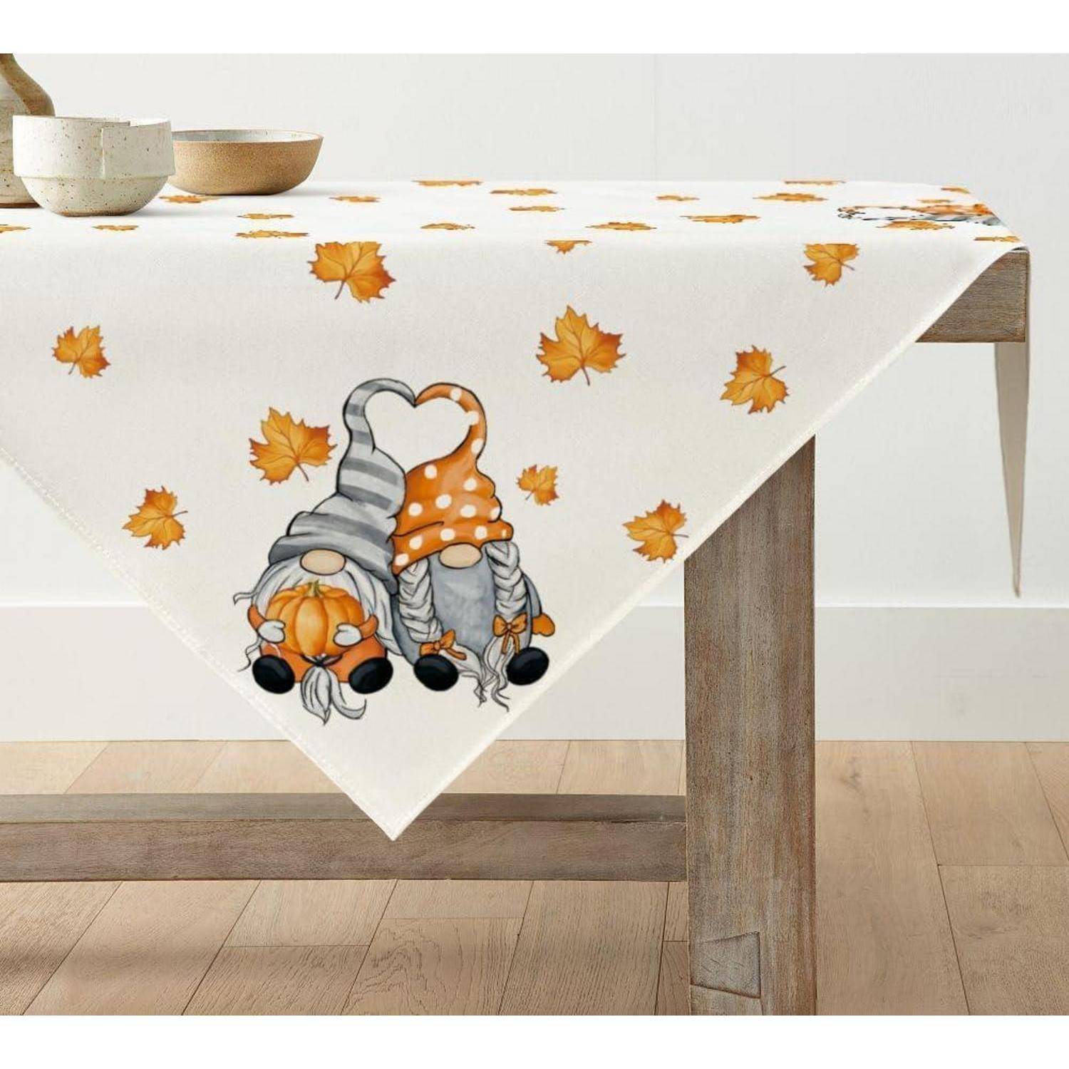 Tisch mit einem weißen Tuch, das Herbst-Illustrationen von Gnomen und orangefarbenen Blättern zeigt, mit einer Holzschüssel oben drauf.