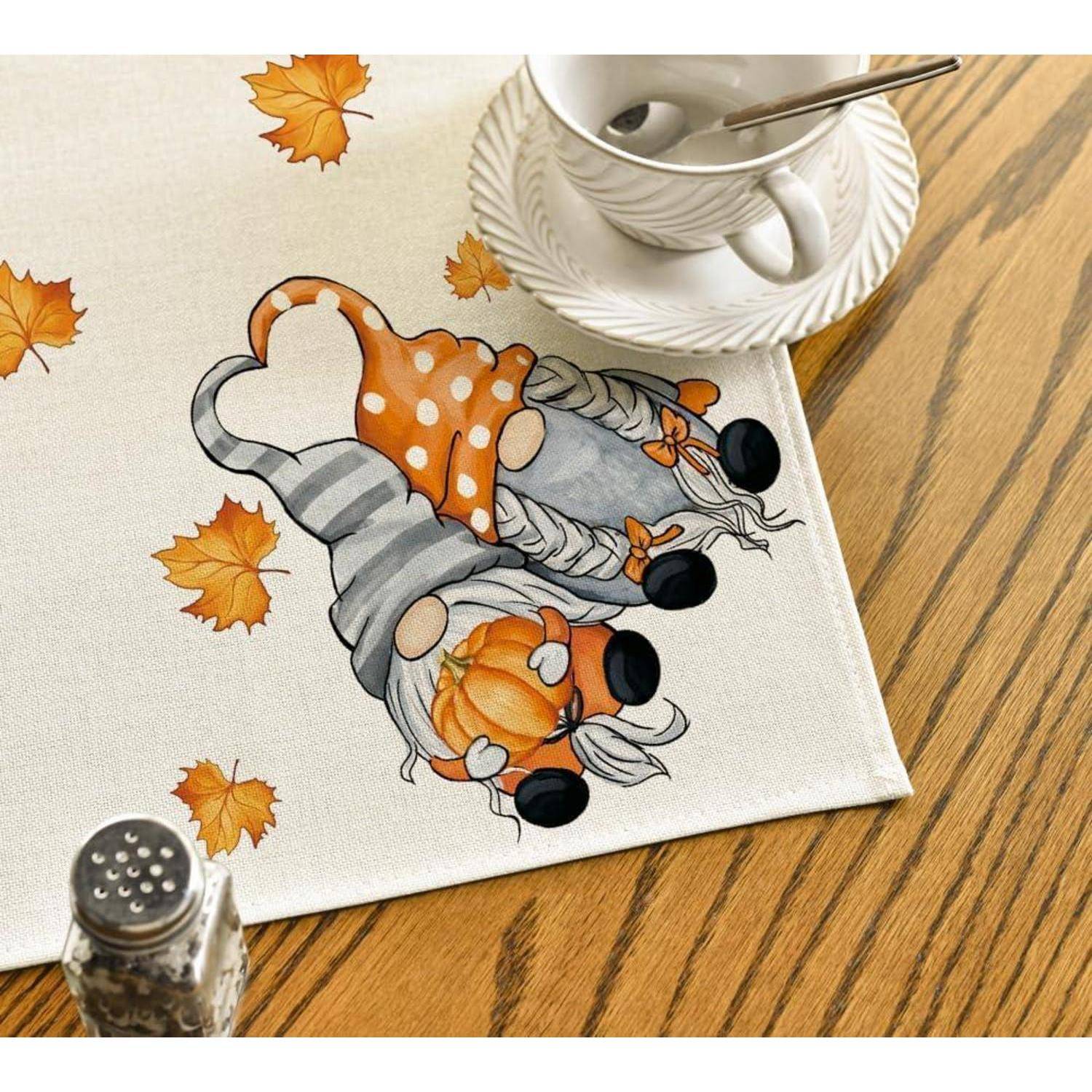 Tischläufer mit Herbstblättern und zwei Gnomen in orangefarbenen und grauen Outfits, in der Nähe von gestapelten weißen Tellern und einer Teetasse auf einem Holztisch.