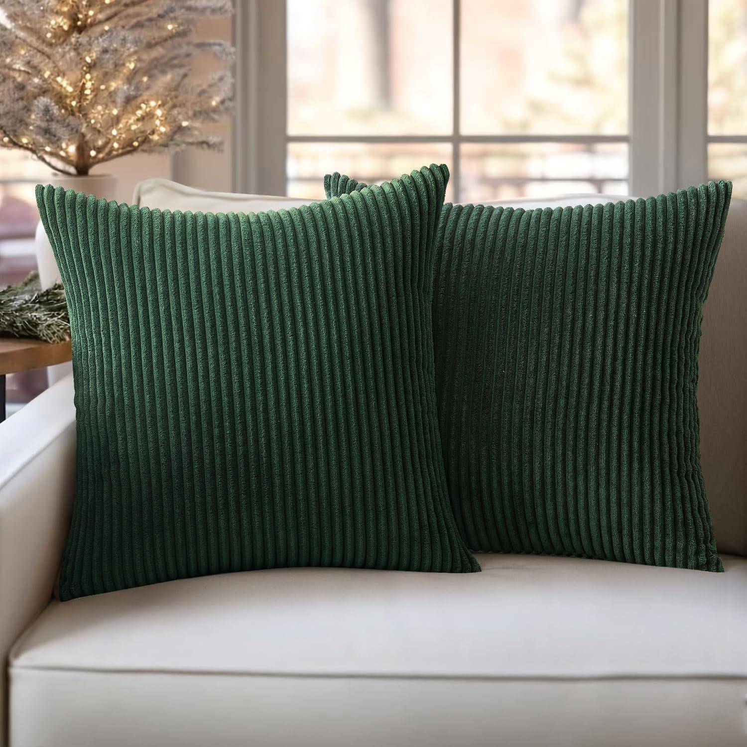 Zwei dunkelgrüne Cordkissen auf einem weißen Sofa, vor einem sanft beleuchteten Fenster und einem dekorierten Baum, die eine gemütliche Atmosphäre schaffen.