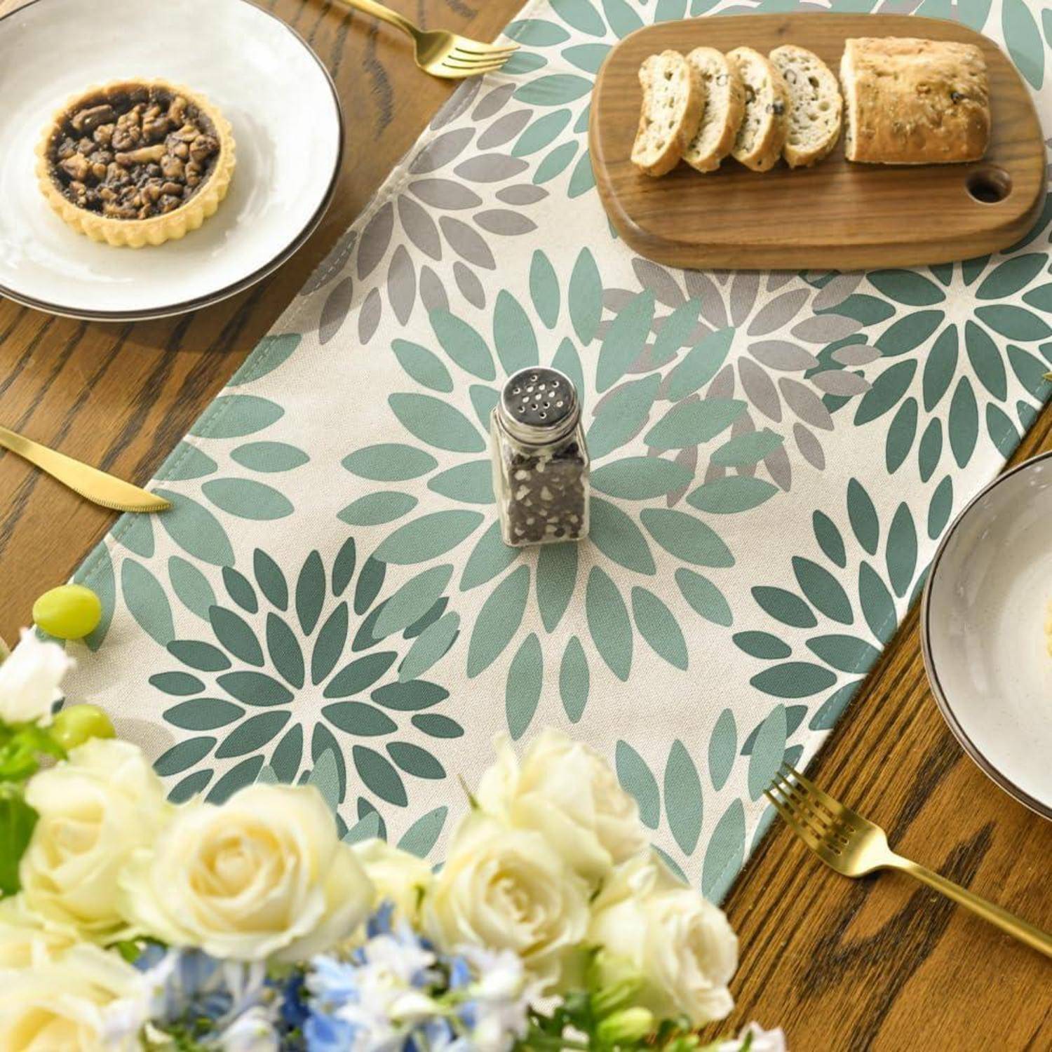 Ein Esstisch, der mit einem blumenmustrigen Läufer gedeckt ist und eine kleine Pastete, in Scheiben geschnittenes Brot auf einem Holzbrett sowie einen Pfefferstreuer zeigt. Weiße und blaue Blumen befinden sich im Vordergrund.