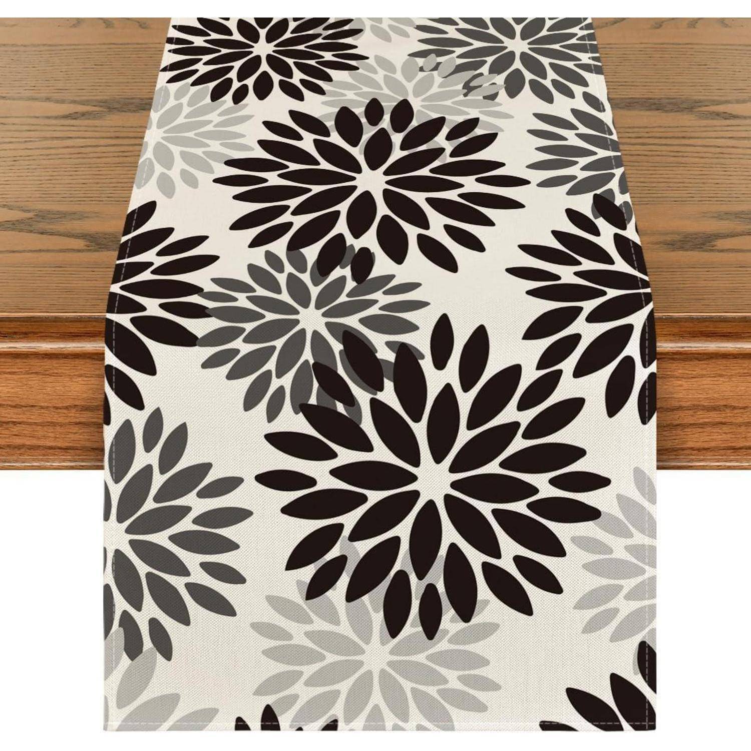 Ein Tischläufer mit einem Blumenmuster in Schwarz- und Grautönen auf beigem Hintergrund, auf einem Holztisch platziert.