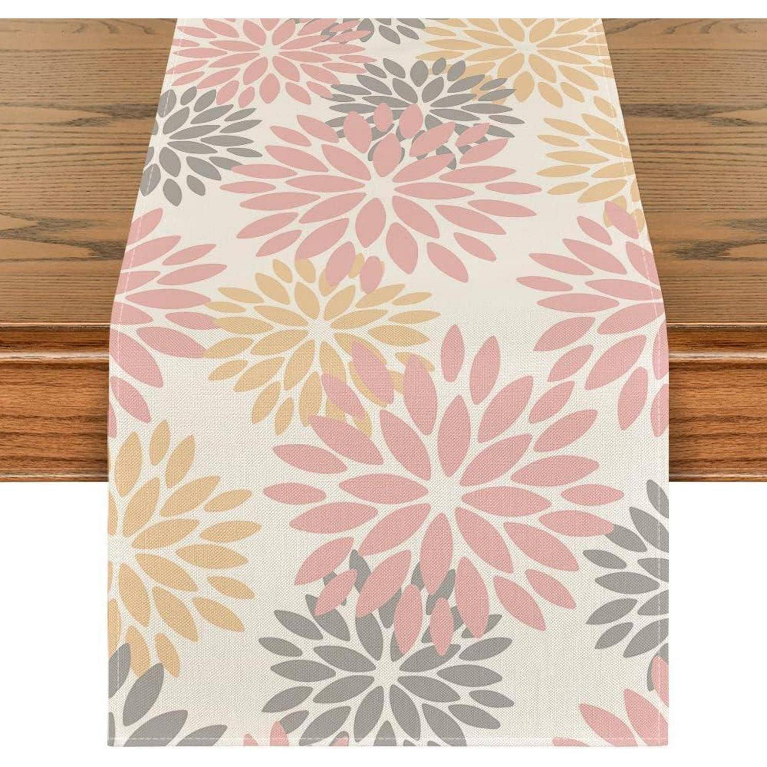 Ein Tischläufer mit einem Blumenmuster, das große rosa, gelbe und graue Blumen auf weißem Hintergrund zeigt, der auf einem Holztisch platziert ist.
