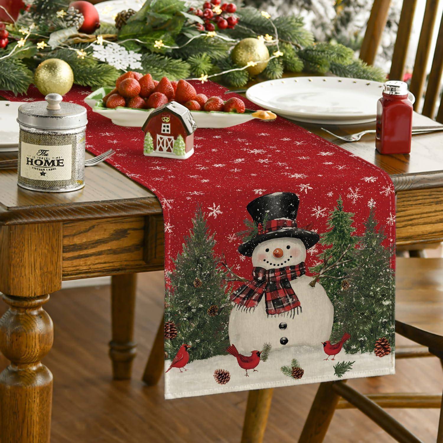 Ein festlich gedeckter Tisch mit einem roten Läufer mit Schneemann-Motiv, umgeben von Weihnachtsdekoration, Erdbeeren, einem Hausornament und einem Marmeladenglas.