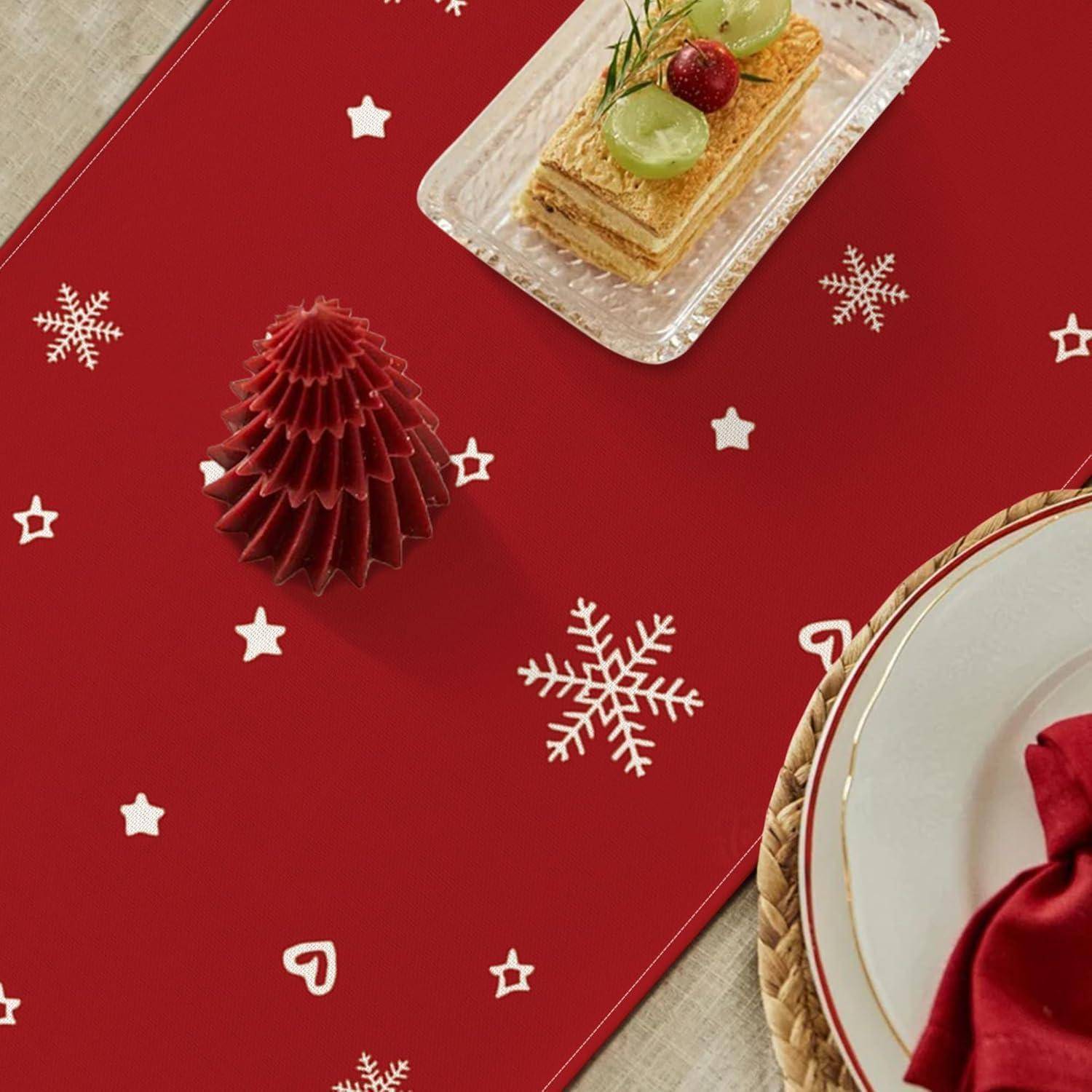 Ein roter Tischläufer mit weißen Schneeflocken und Sternen schmückt einen Tisch, auf dem eine Feiertagskarte, ein Dessertteller mit Trauben und einer Kirsche sowie ein halb sichtbarer Essteller platziert sind.