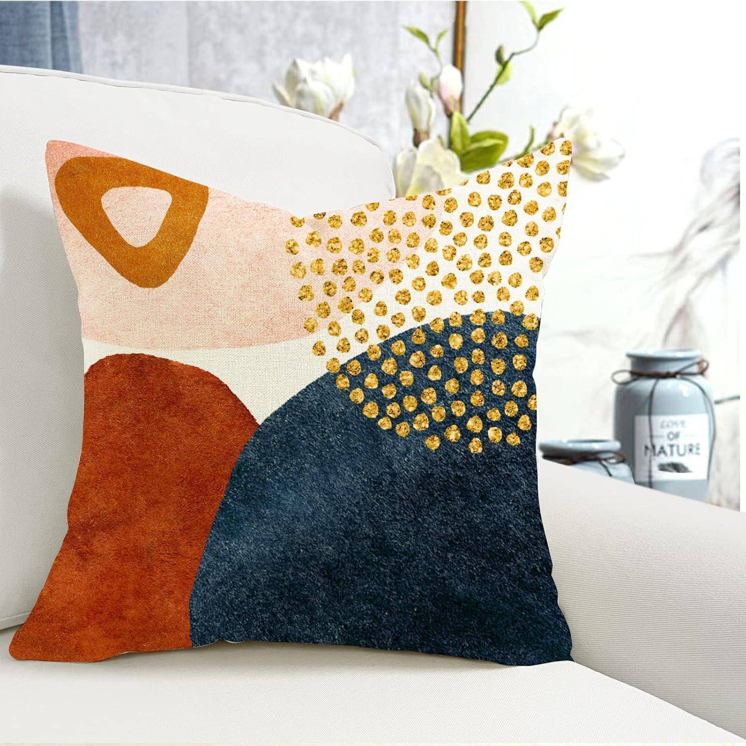 Ein dekorativer Kissenbezug mit abstrakten geometrischen Mustern in Orange, Rosa, Marineblau und Gold auf einem weißen Stuhl, mit einer Pflanze im Hintergrund.
