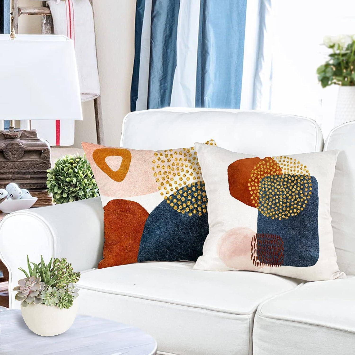 Ein weißes Sofa mit zwei dekorativen Kissen, die abstrakte geometrische Muster in Orange-, Blau- und Goldtönen aufweisen. Eine Lampe und eine Zimmerpflanze befinden sich in der Nähe.