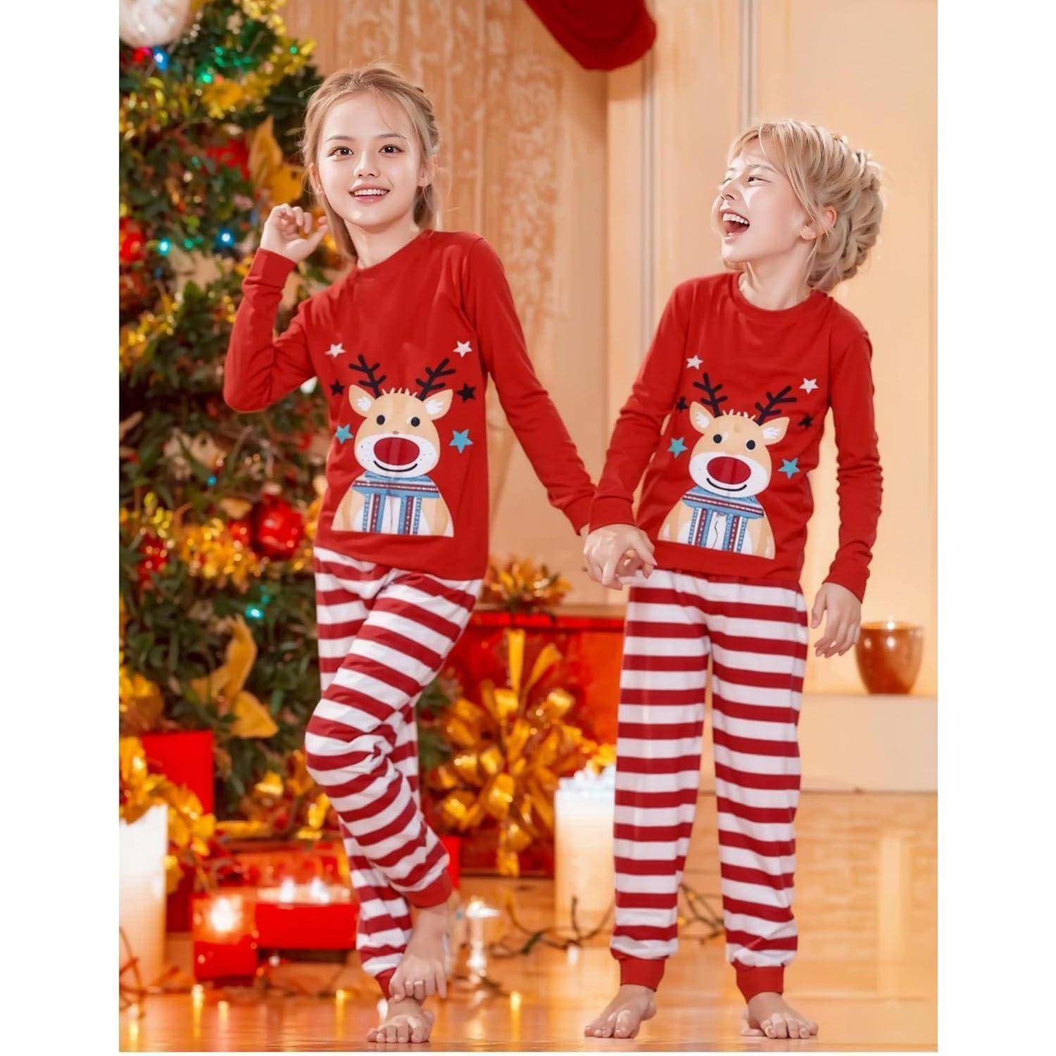 Zwei Kinder in passenden roten Rentier-Pyjamas halten sich an den Händen und lächeln vor einem geschmückten Weihnachtsbaum mit bunten Christbaumkugeln.