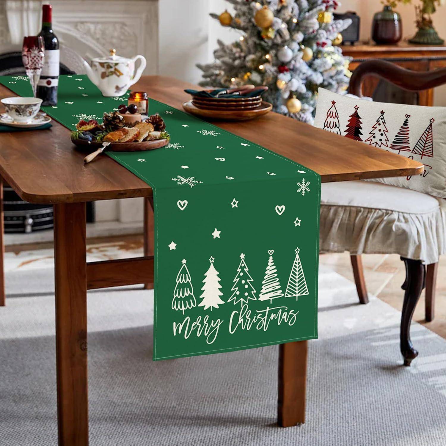 Ein Esstisch, der für Weihnachten gedeckt ist, mit einem grünen Tischläufer, der weiße Bäume und den Text ‚Frohe Weihnachten' trägt. Die Stühle haben festliche Designs.