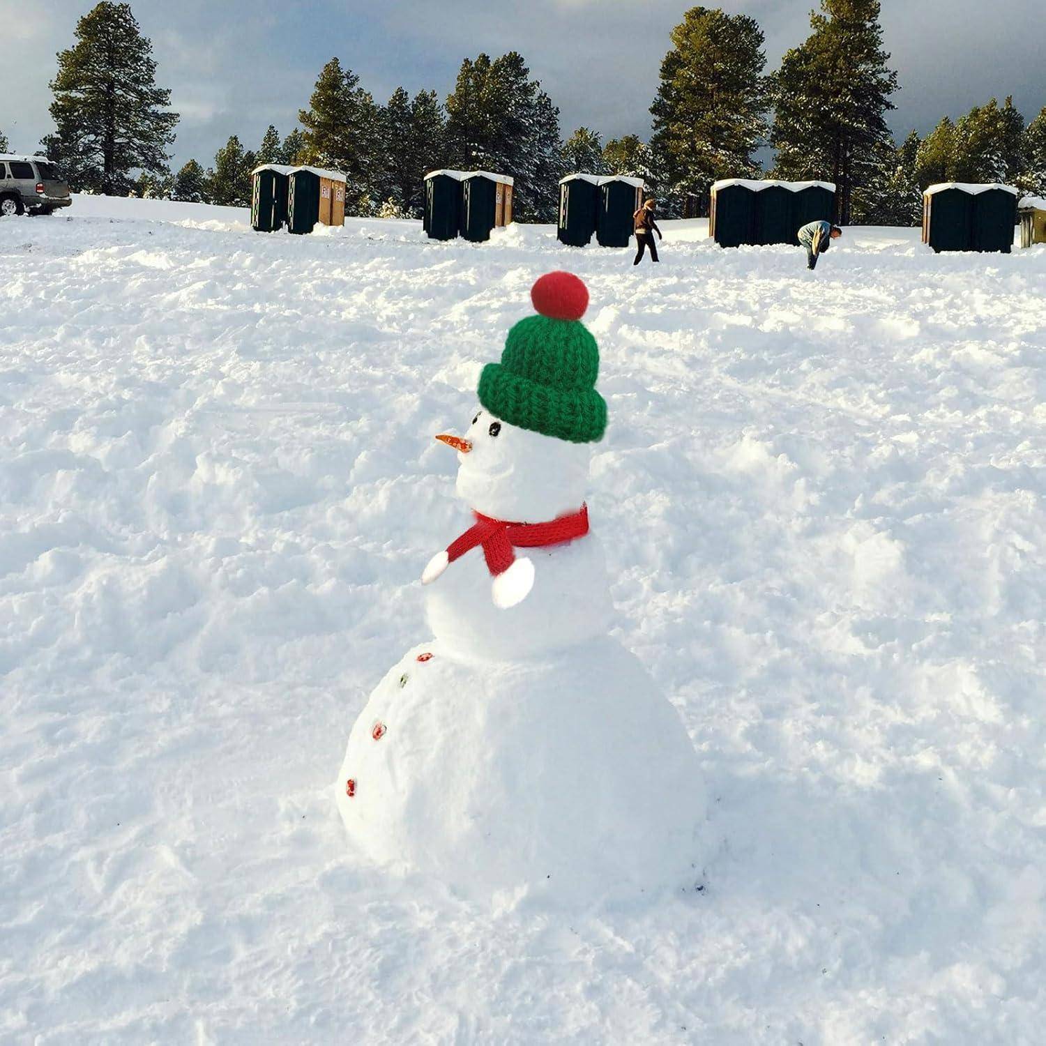 Ein Schneemann mit einem grünen Hut und einem roten Schal steht auf schneebedecktem Boden, mit Bäumen und mobilen Toiletten im Hintergrund.