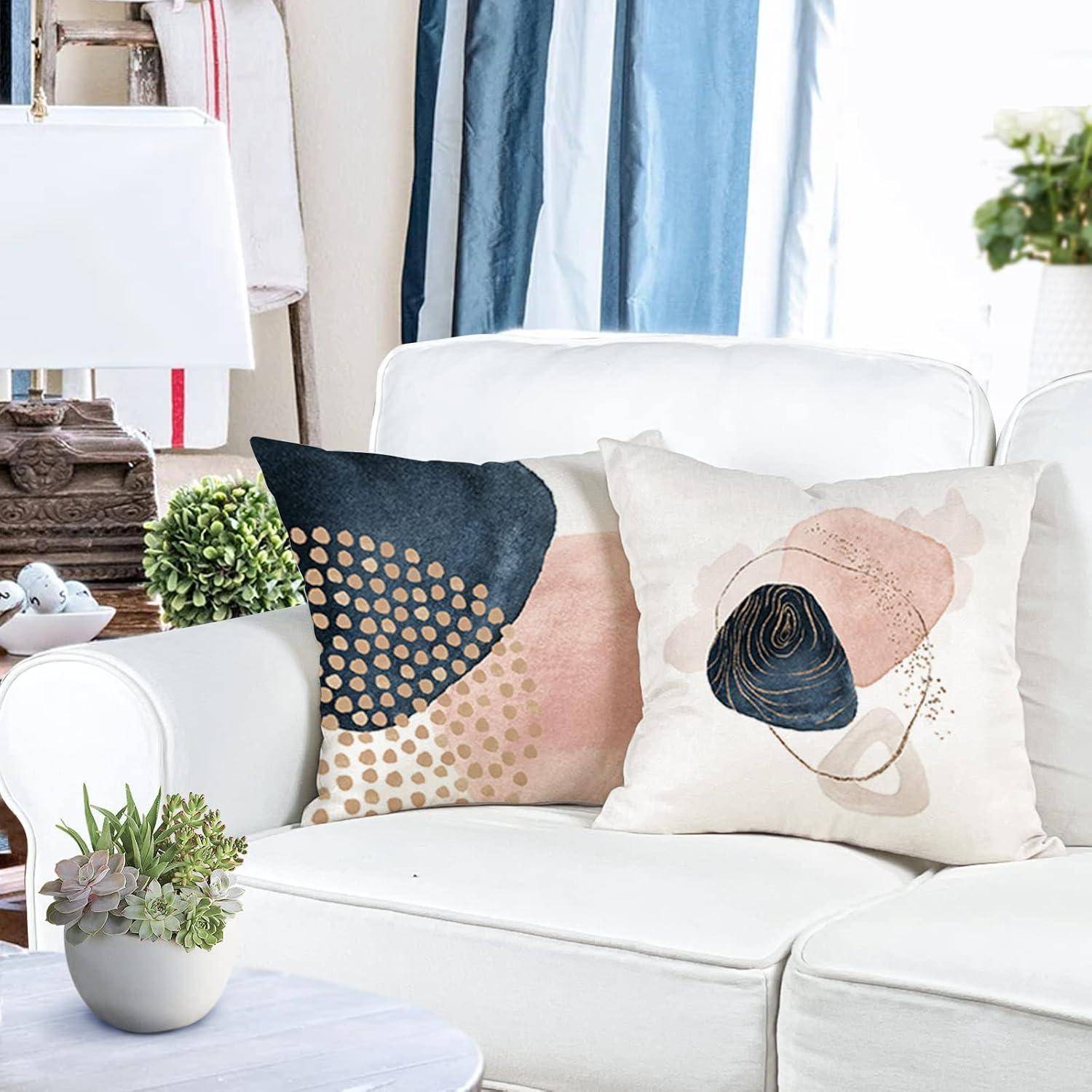 Ein gemütliches Wohnzimmer mit einem weißen Sofa, geschmückt mit abstrakten gemusterten Kissen in Blau-, Beige- und Rosatönen, neben einer Lampe und Pflanzen.