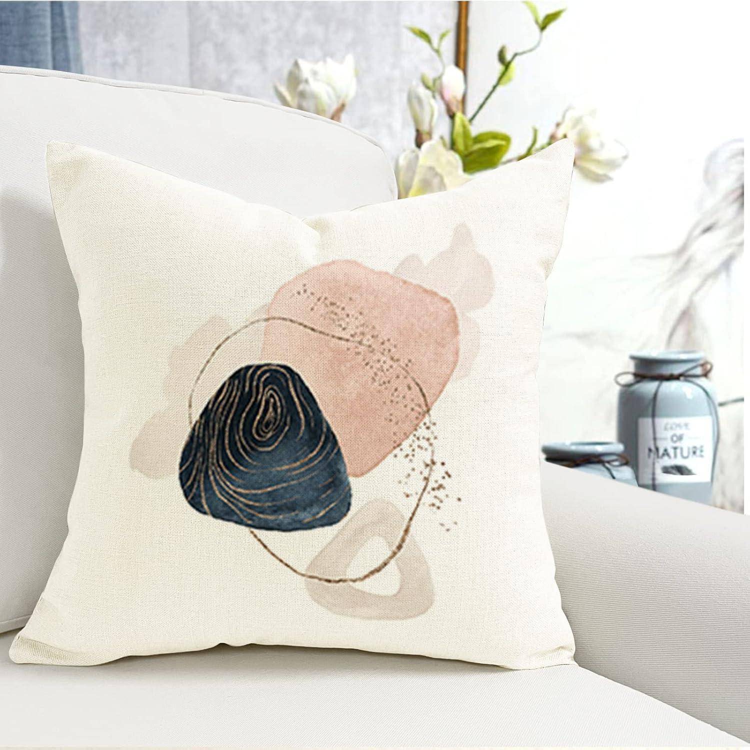 Ein dekorativer Kissenbezug mit abstrakten Formen in Beige, Rosa und Marineblau liegt auf einem weißen Sofa. Blumen und eine Kerze sind im Hintergrund unscharf.