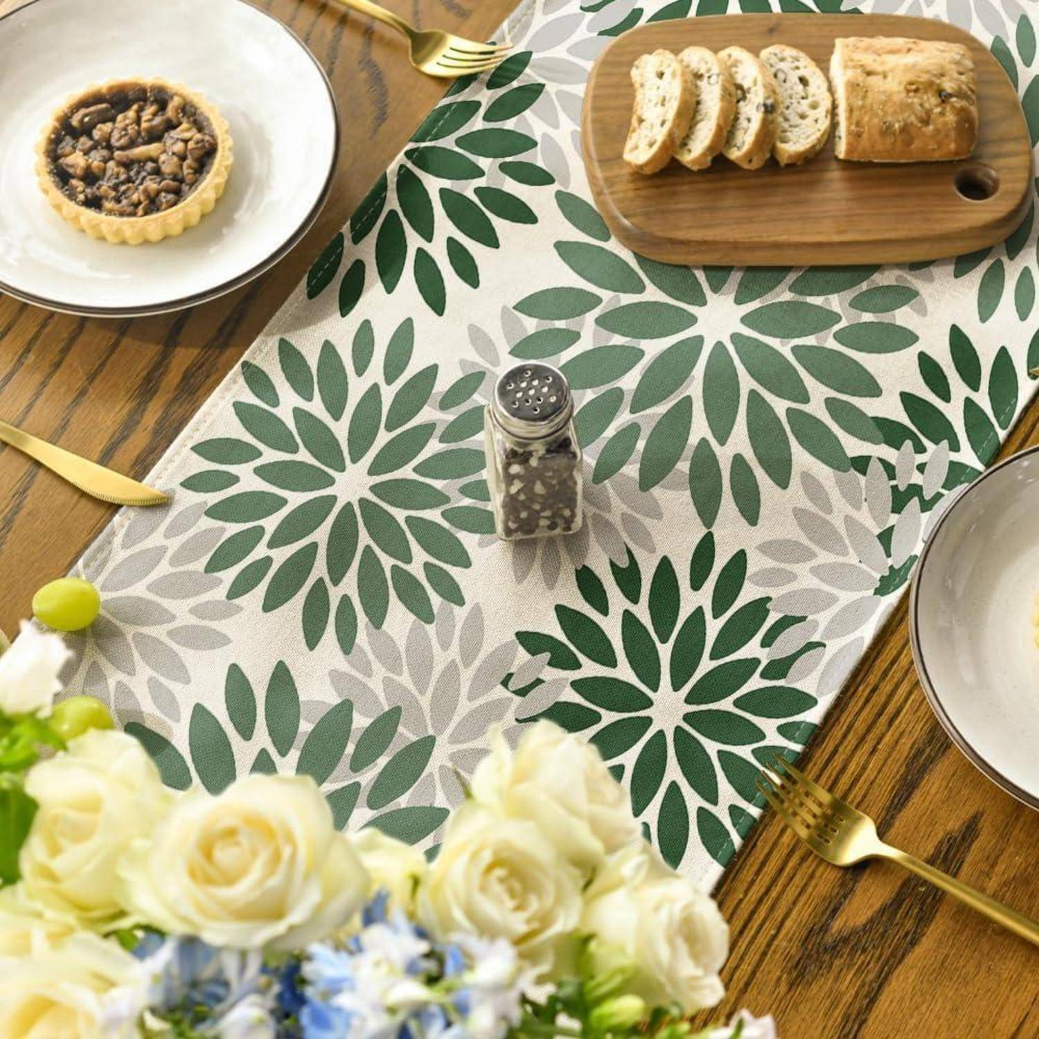 Esstisch, der mit einem blumenmustrigen Läufer gedeckt ist, mit Brot auf einem Holzbrett, einem kleinen Törtchen, einem Pfefferstreuer und einer Blumenvase.