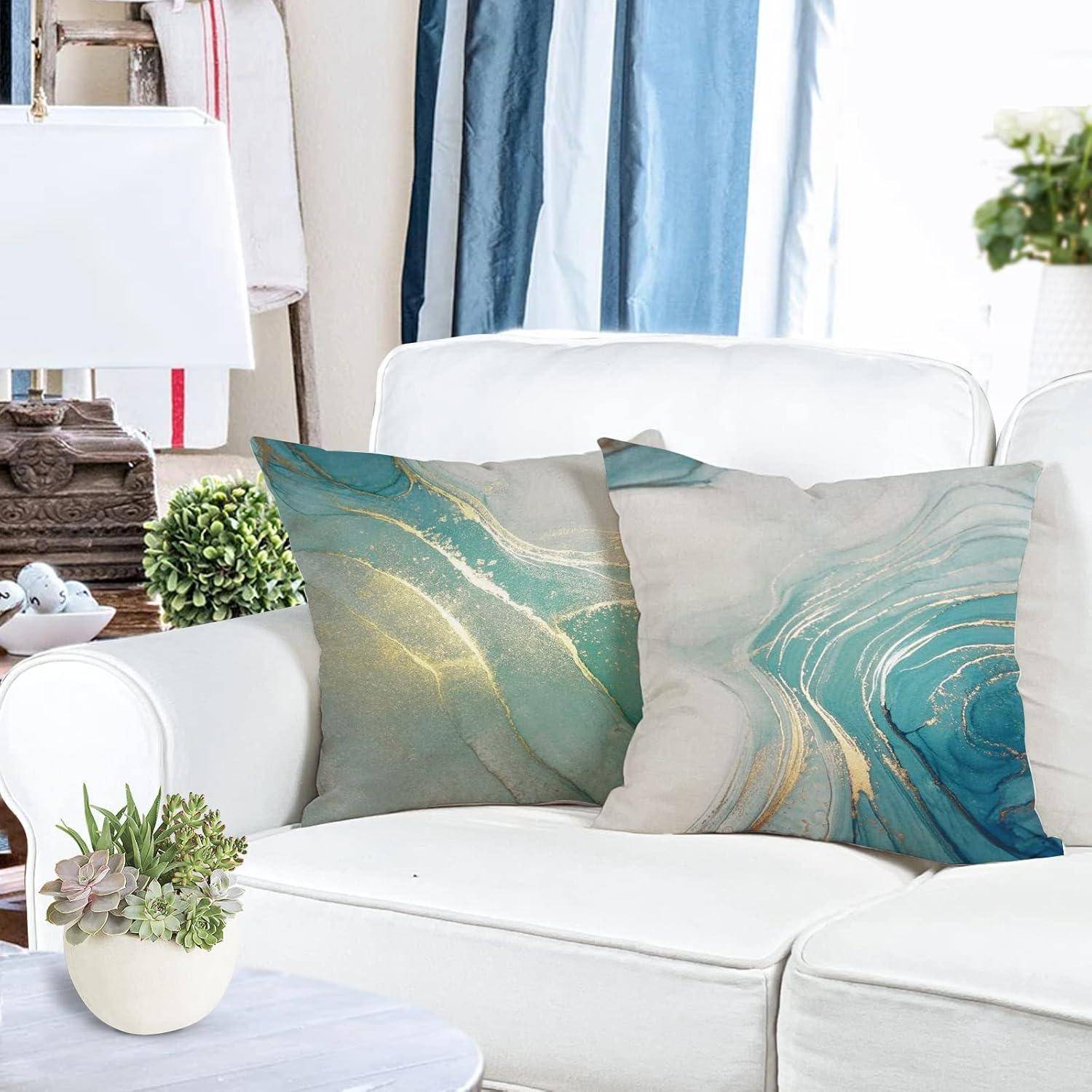 Ein weißes Wohnzimmer mit einem Sofa und zwei dekorativen Kissen mit abstrakten, geschwungenen grünen und beigen Mustern. Eine Lampe, Bücher und Pflanzen befinden sich in der Nähe.