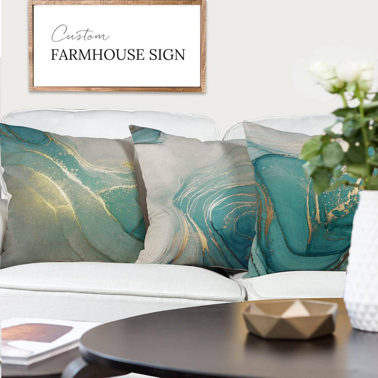 Ein weißes Sofa mit abstrakten Teal- und Goldkissen steht unter einem gerahmten Schild mit der Aufschrift ‚Custom Farmhouse Sign', neben einer Vase mit Blumen.