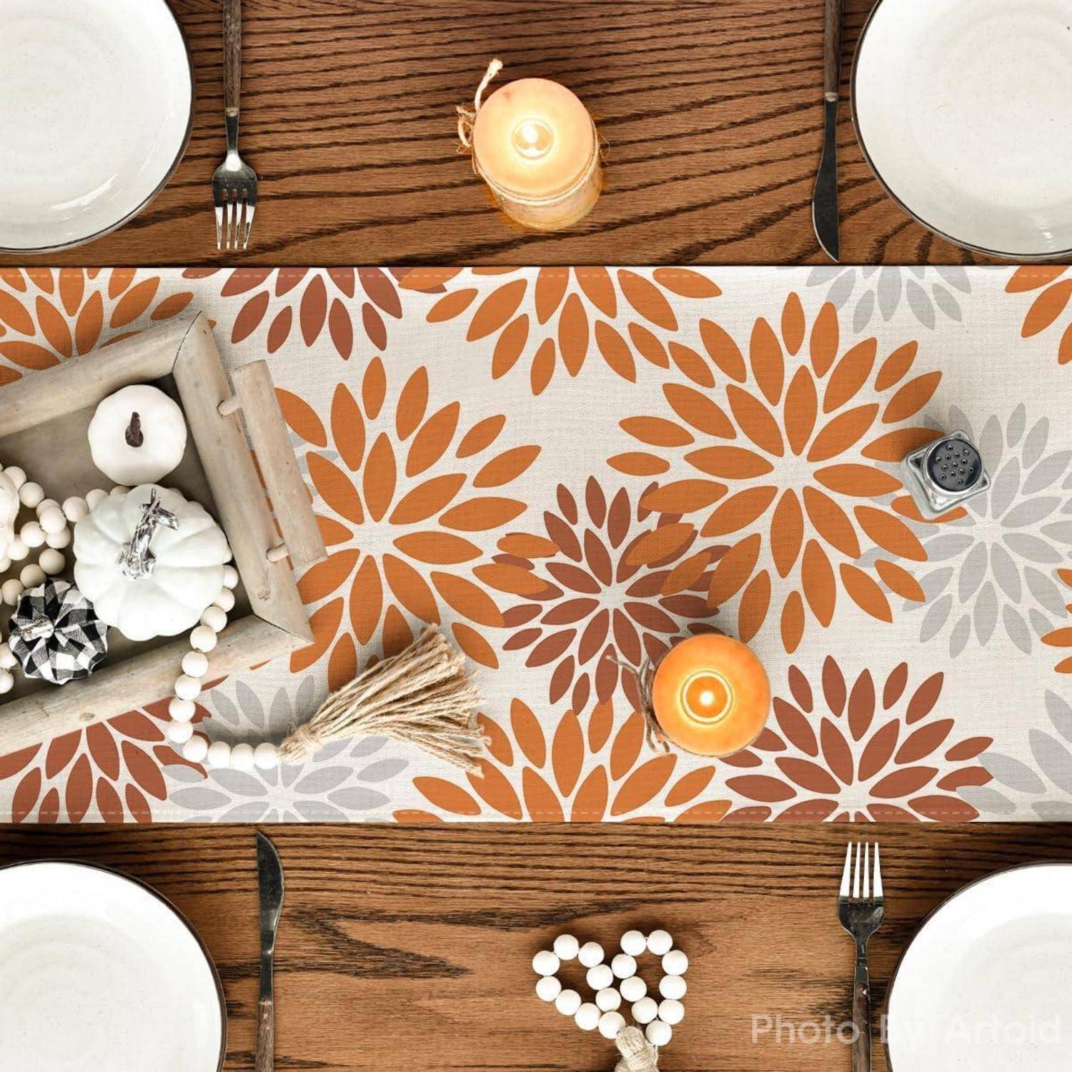 Ein für eine Mahlzeit gedeckter Tisch mit weißen Tellern, schwarzen Gabeln und einem dekorativen, herbstlich gestalteten Blumen-Tischläufer. Kerzen und ein Mittelstück schmücken den Tisch.