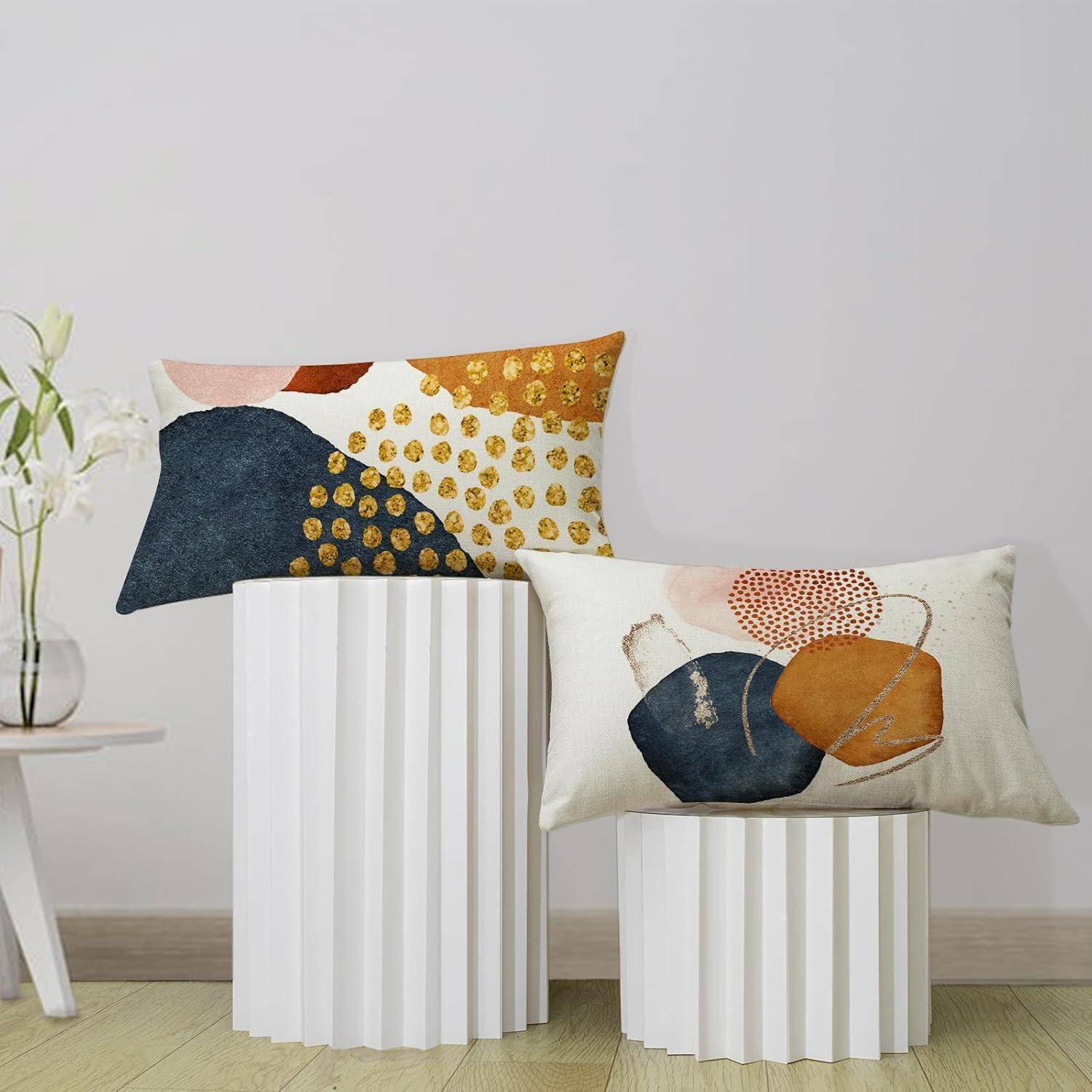 Zwei dekorative Kissen mit abstrakten Mustern auf zylindrischen weißen Ständern, neben einem kleinen Tisch mit weißen Blumen, in einem minimalistischen Raum.