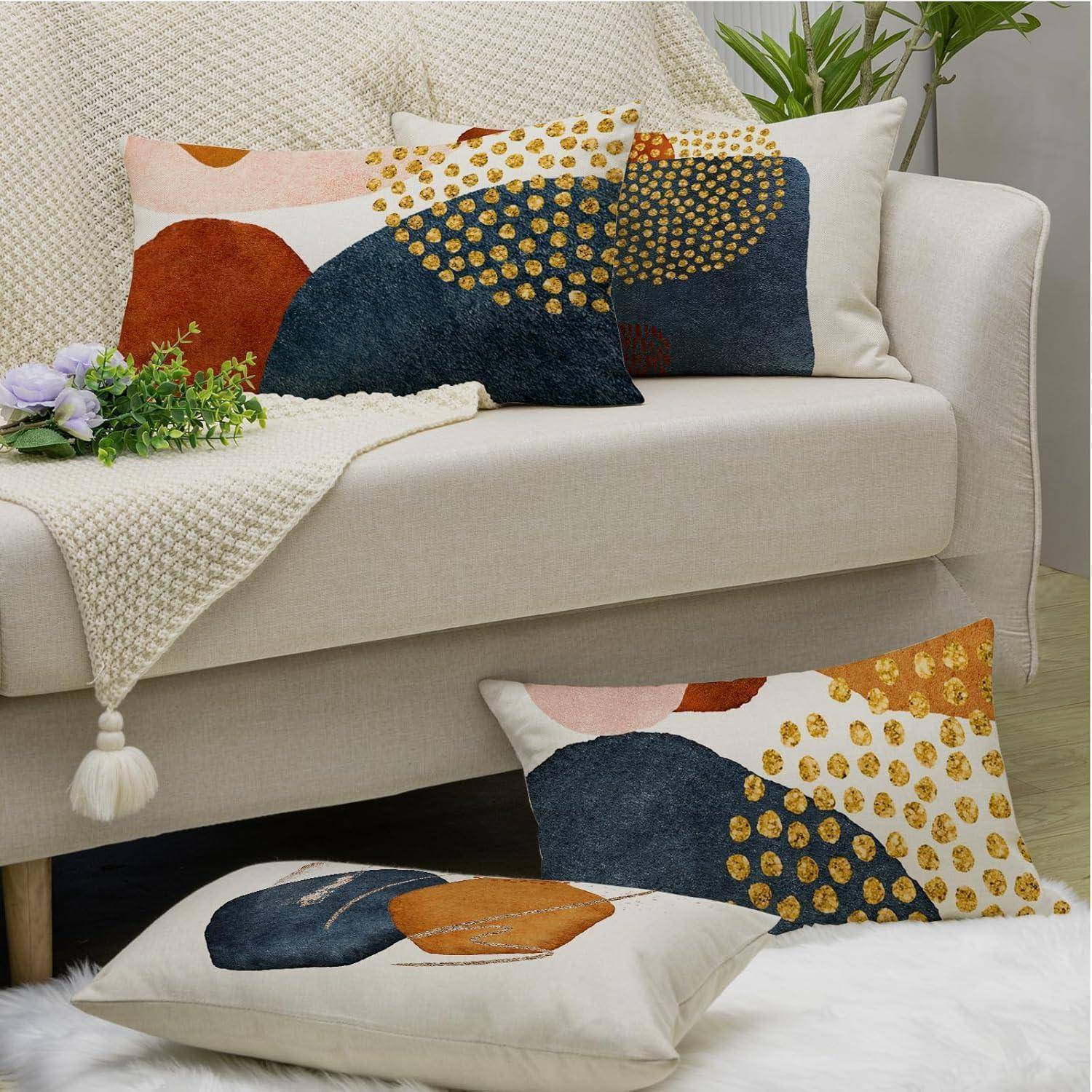 Ein beiger Sofa mit geometrisch gemusterten Kissen, die Orange-, Marineblau- und Golddesigns aufweisen. Eine cremefarbene gestrickte Decke und eine Zimmerpflanze vervollständigen die Dekoration.