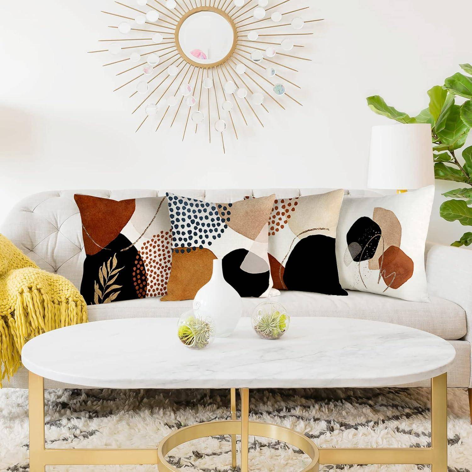 Ein modernes Wohnzimmer mit einem weißen Sofa, das mit abstrakt gemusterten Kissen versehen ist, einem runden Marmor-Couchtisch mit Deko und einem Sonnenspiegel an der Wand.
