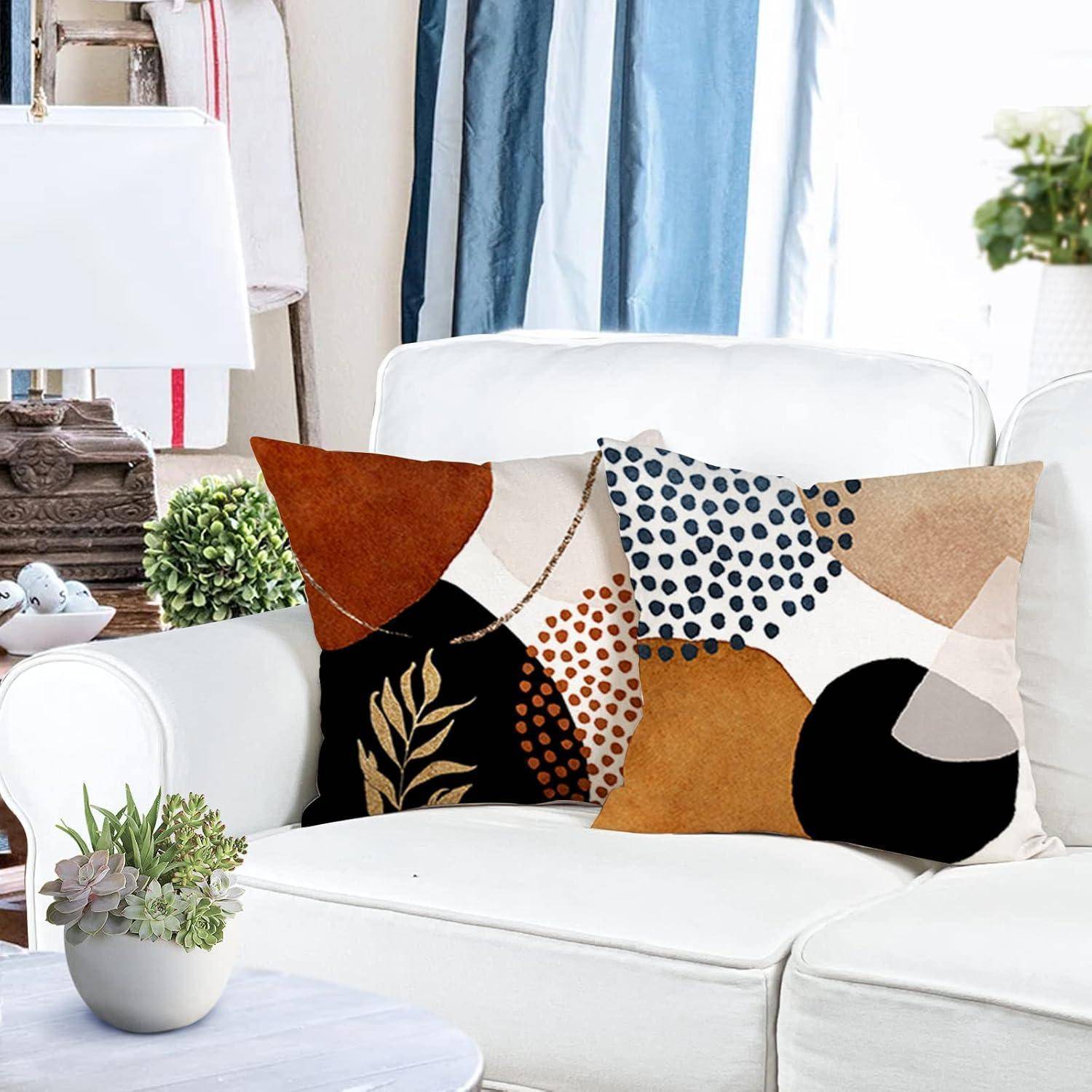Ein weißer Sessel mit abstrakten Kissen in erdigen Tönen steht in einem gemütlichen Wohnzimmer. Eine Pflanze und eine Lampe befinden sich in der Nähe und vervollständigen die Einrichtung.