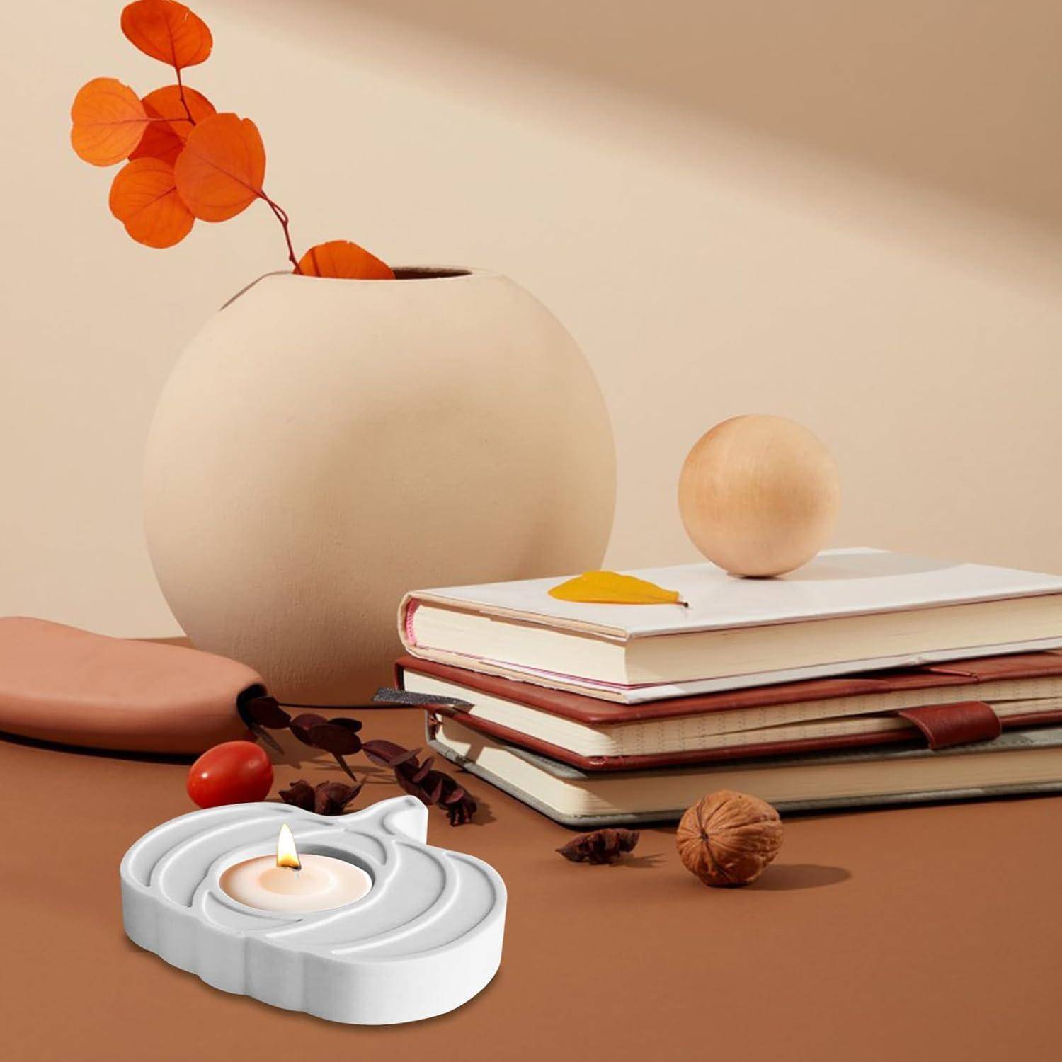 Ein weißer Kerzenhalter mit einer brennenden Kerze, Bücher gestapelt neben einer hellbraunen Vase mit orangefarbenen Blättern, auf einer beigen Oberfläche.