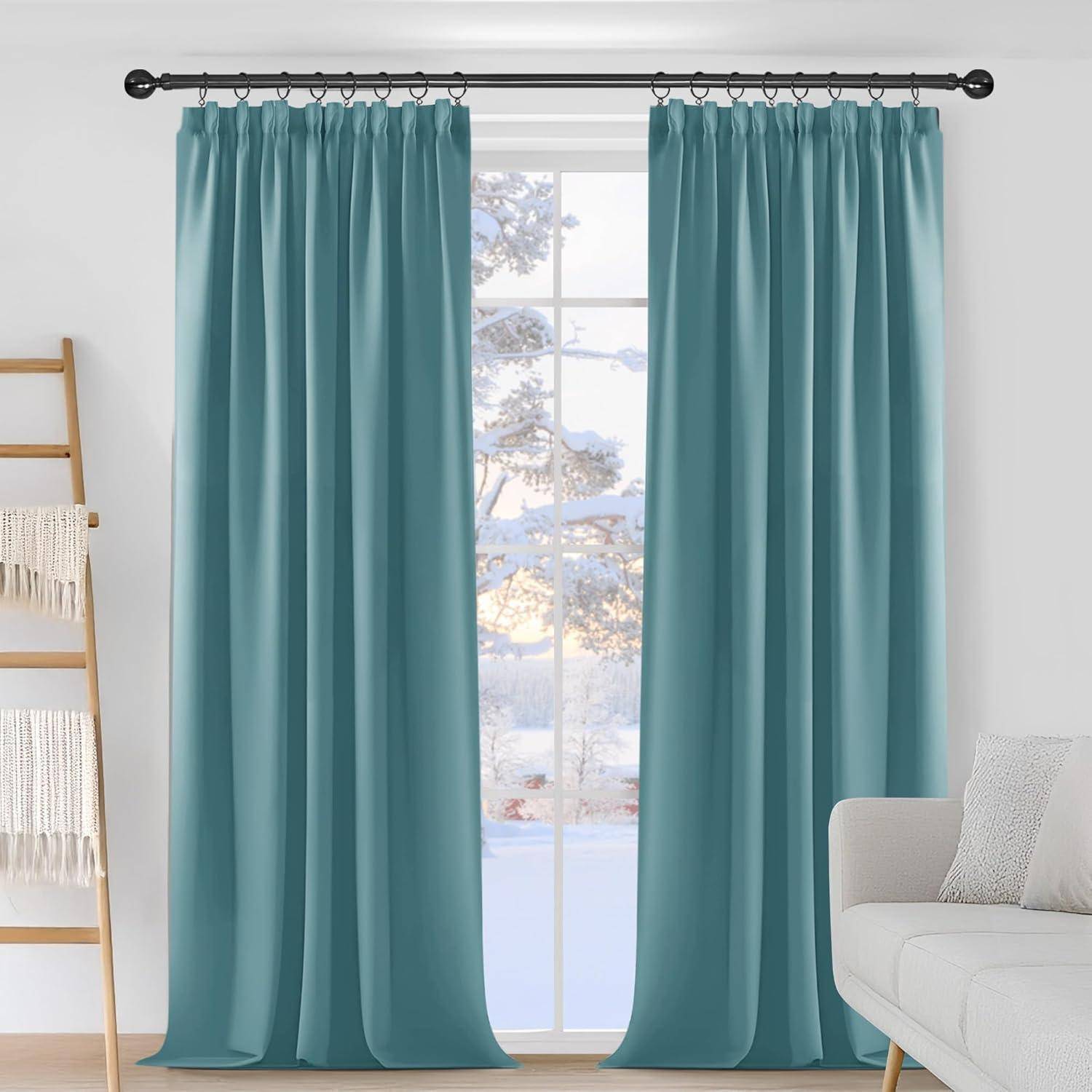 Ein Paar Petrol-Vorhänge hängt an einer schwarzen Stange vor einem Fenster mit einer verschneiten Landschaft dahinter; daneben stehen ein weißes Sofa und ein Leiterregal.