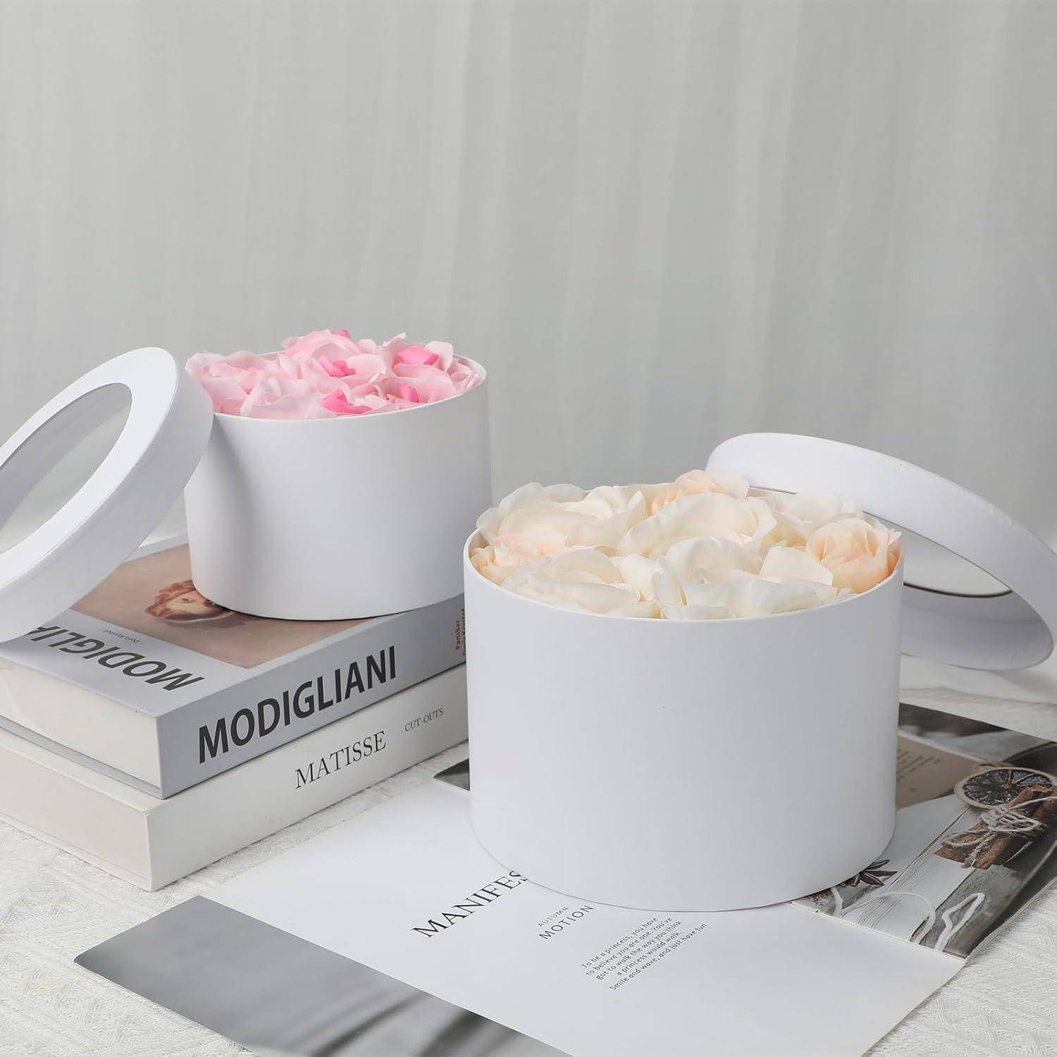 Zwei runde weiße Schachteln, eine mit rosa Blütenblättern und die andere mit cremefarbenen Blütenblättern, liegen auf Büchern mit den Titeln 