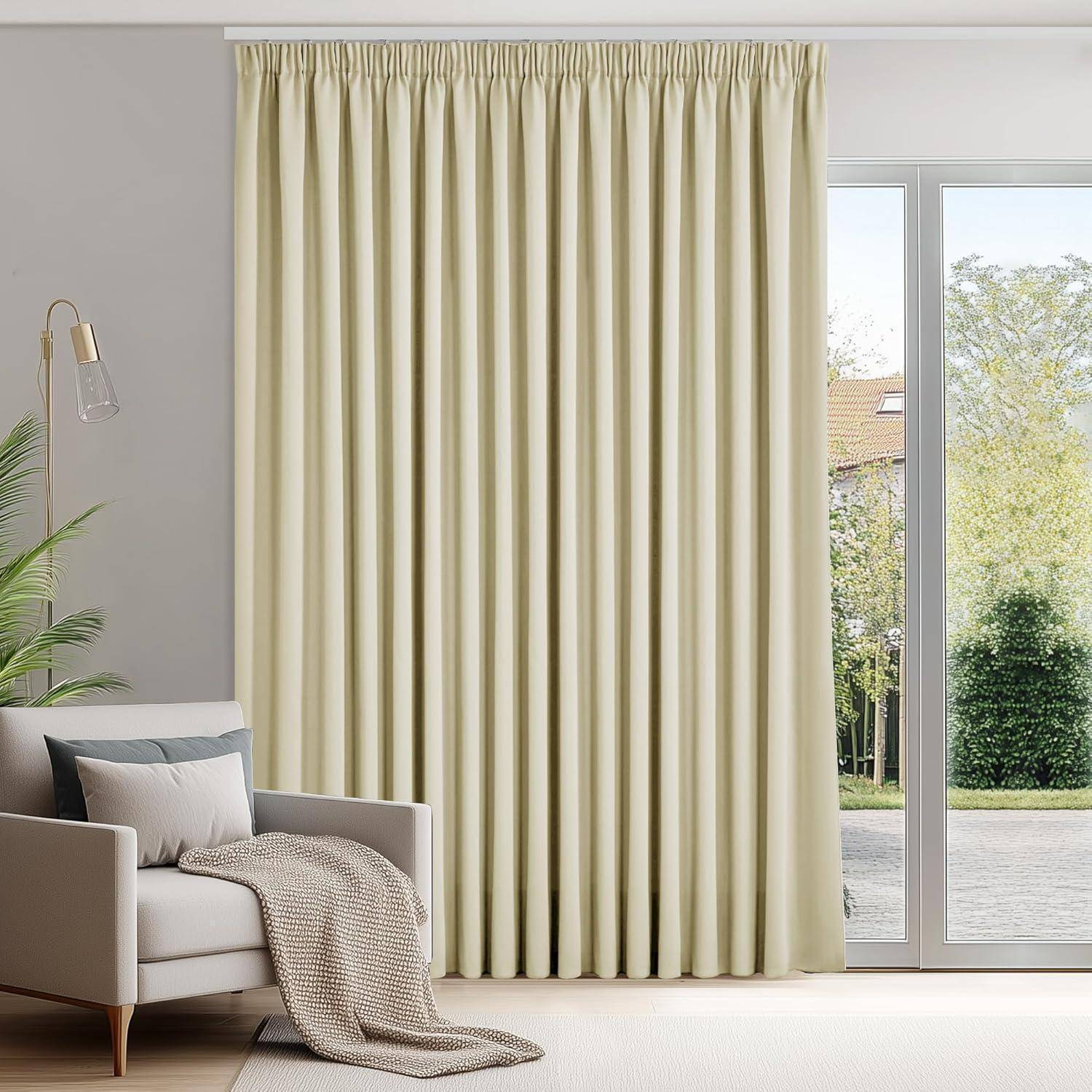 Ein Wohnzimmer mit einem beigen Sofa, einem Kissen und einer grauen Decke. Cremefarbene Vorhänge verdecken ein großes Fenster, hinter dem Grünpflanzen zu sehen sind.