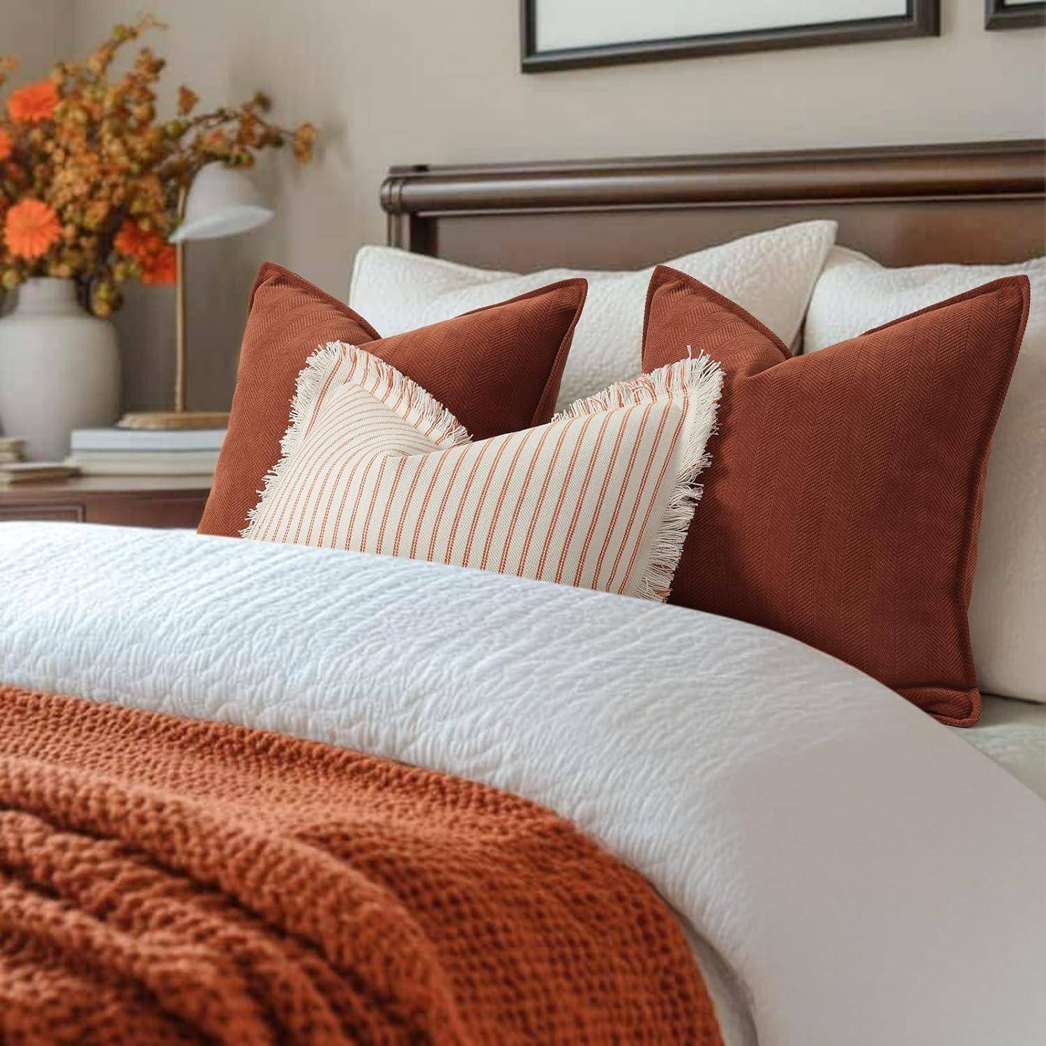 Ein sauber gemachtes Bett mit weißen und rostfarbenen Kissen und einer passenden Decke, eingebettet in ein gemütliches Schlafzimmer mit warmtöniger Einrichtung und Blumen.
