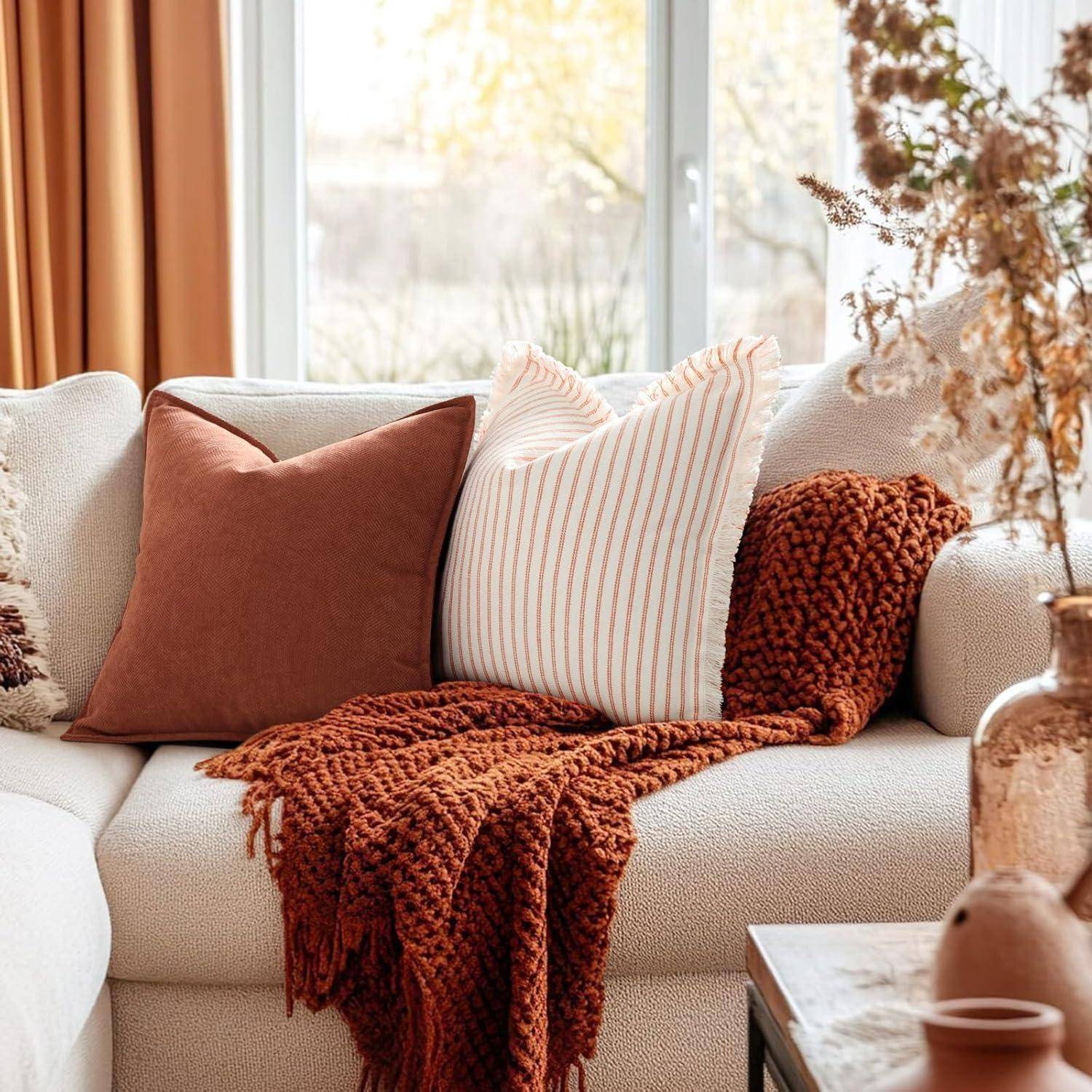 Eine gemütliche Wohnzimmerszene mit einem beigen Sofa, das mit braunen und gestreiften Kissen und einem strukturierten orangefarbenen Überwurf ausgestattet ist, in der Nähe eines großen Fensters.