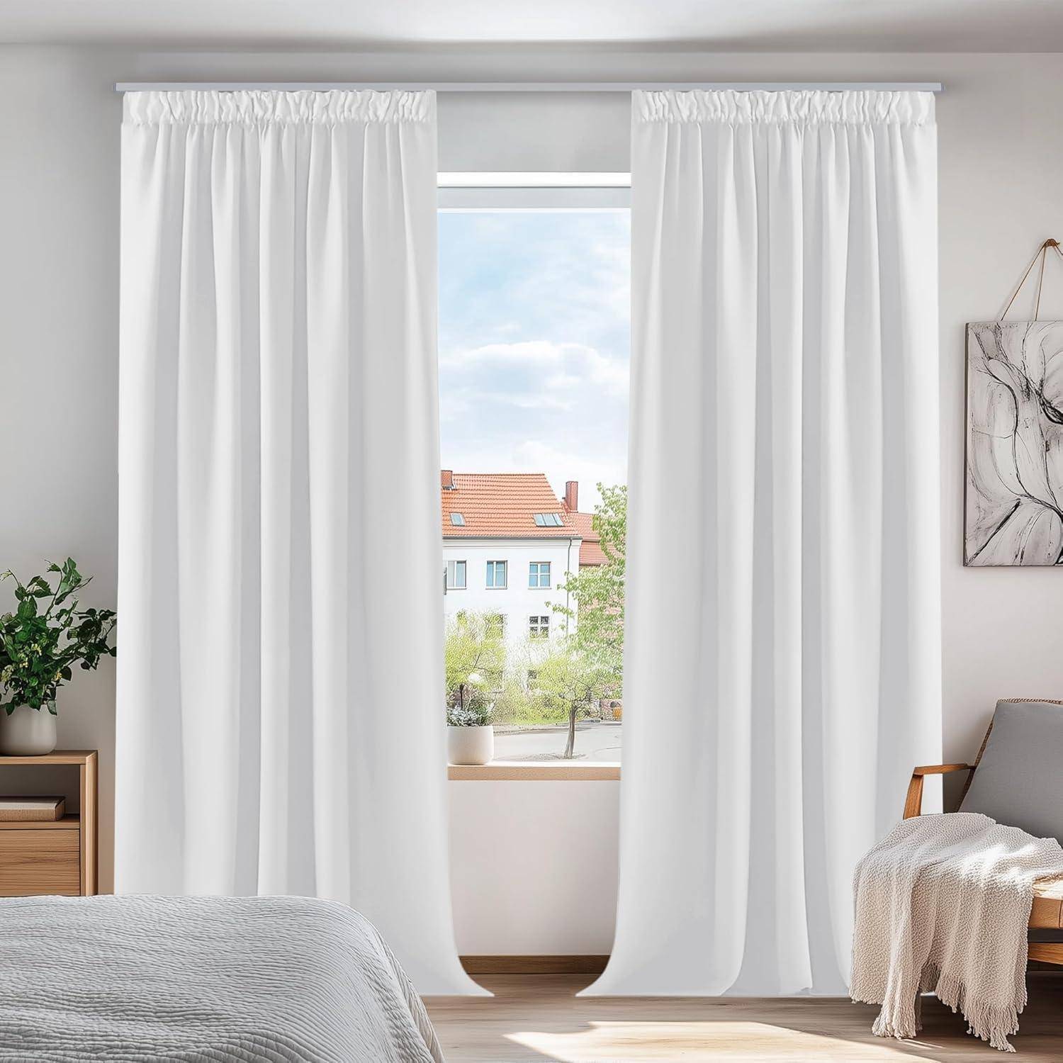 Ein Schlafzimmer mit weißen Vorhängen, die ein Fenster rahmen, das einen Blick auf Häuser und blauen Himmel zeigt. Ein Bett, eine Topfpflanze und Kunstwerke sind sichtbar.