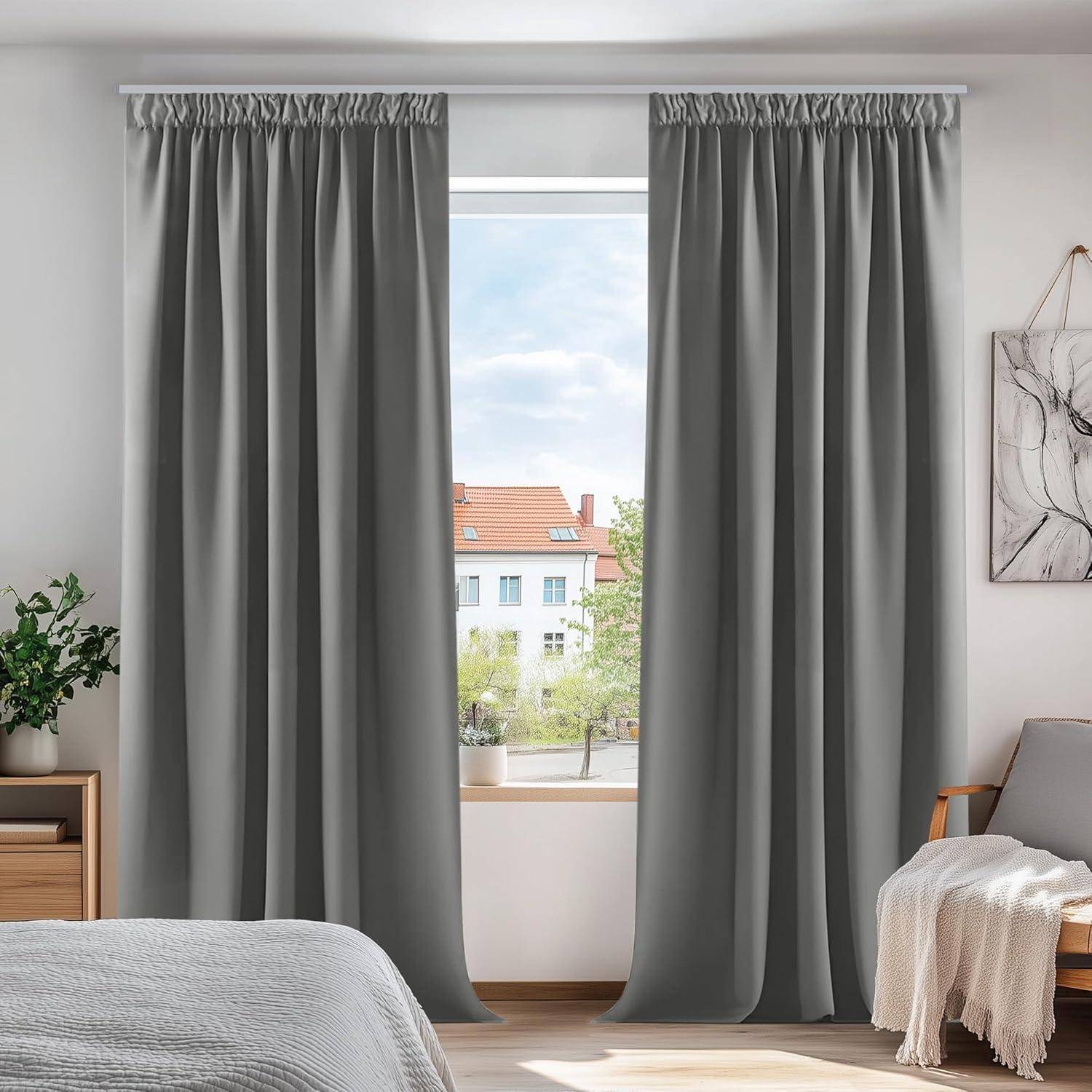 Zimmer mit einem großen Fenster, an dem graue Vorhänge hängen. Eine kleine Topfpflanze steht auf einer Holzkommode. Vom Fenster aus ist ein Blick auf Dächer unter einem wolkenverhangenen Himmel zu sehen.