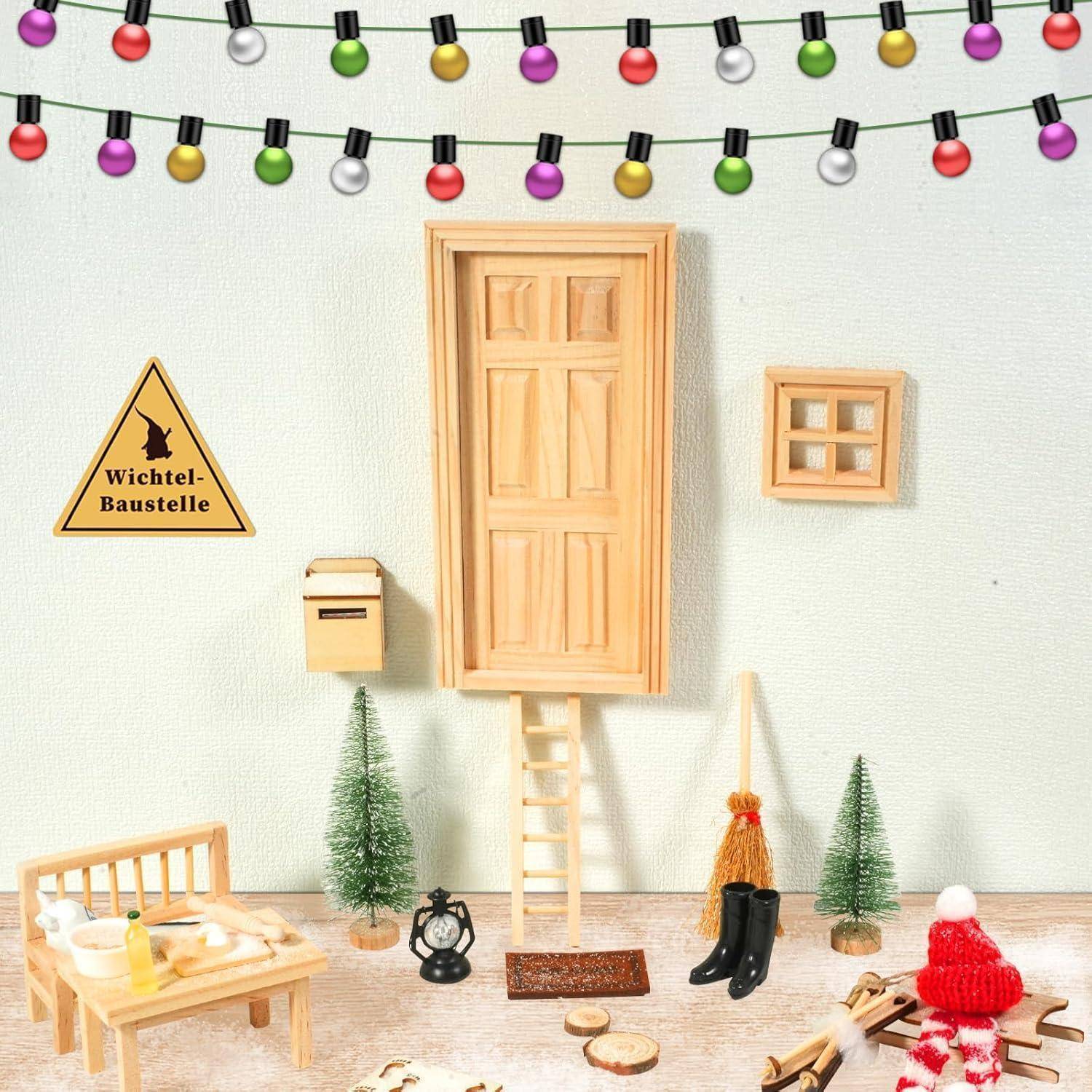 Ein Miniaturaufbau zeigt eine Tür an einer Wand mit einer Leiter darunter, umgeben von kleiner Möblierung, Bäumen und Lichterketten, wodurch eine festliche Szene entsteht.