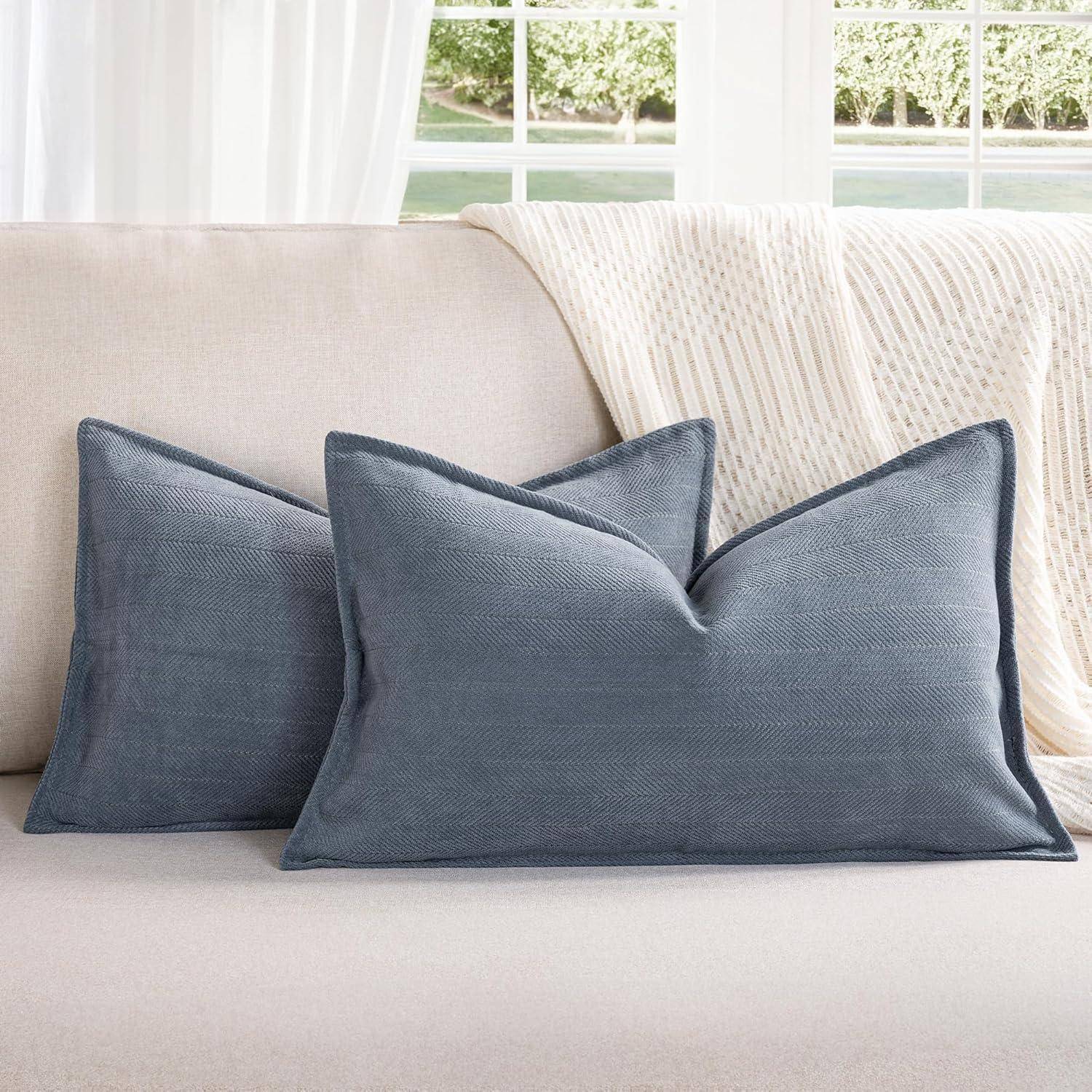 Ein gemütliches Wohnzimmer mit zwei blauen Zierkissen auf einem hellfarbenen Sofa, einer weißen gestrickten Decke und großen Fenstern im Hintergrund.