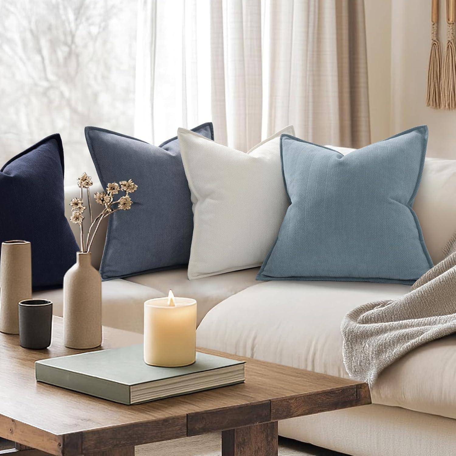 Ein gemütliches Wohnzimmer mit einem weißen Sofa, blauen und grauen Kissen, einem Buch und einer brennenden Kerze auf einem Holzcouchtisch und Blick auf eine Winterlandschaft.