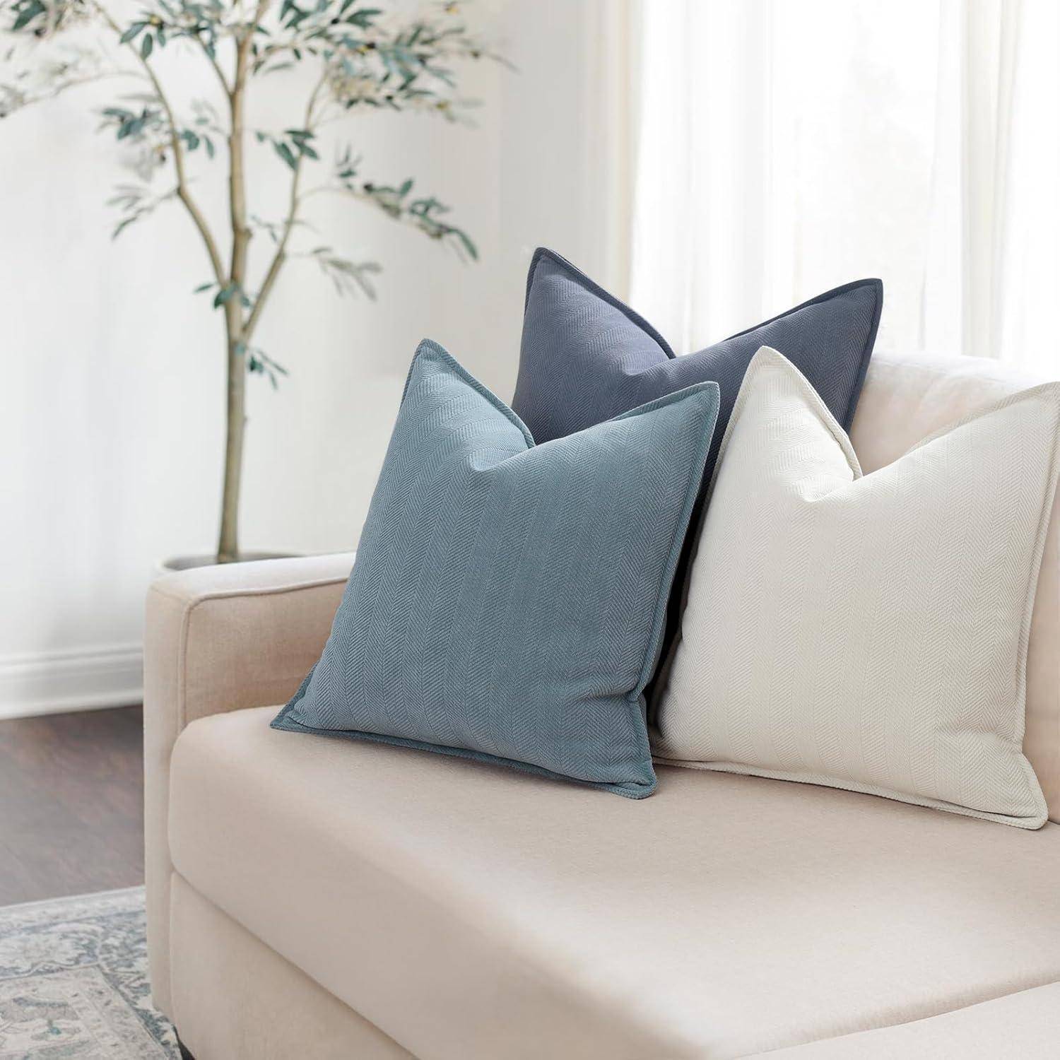 Eine beige Couch mit drei dekorativen Kissen in Blau, Dunkelgrau und Weiß vor einem hellen Hintergrund mit einem Baum, der eine ruhige Atmosphäre schafft.