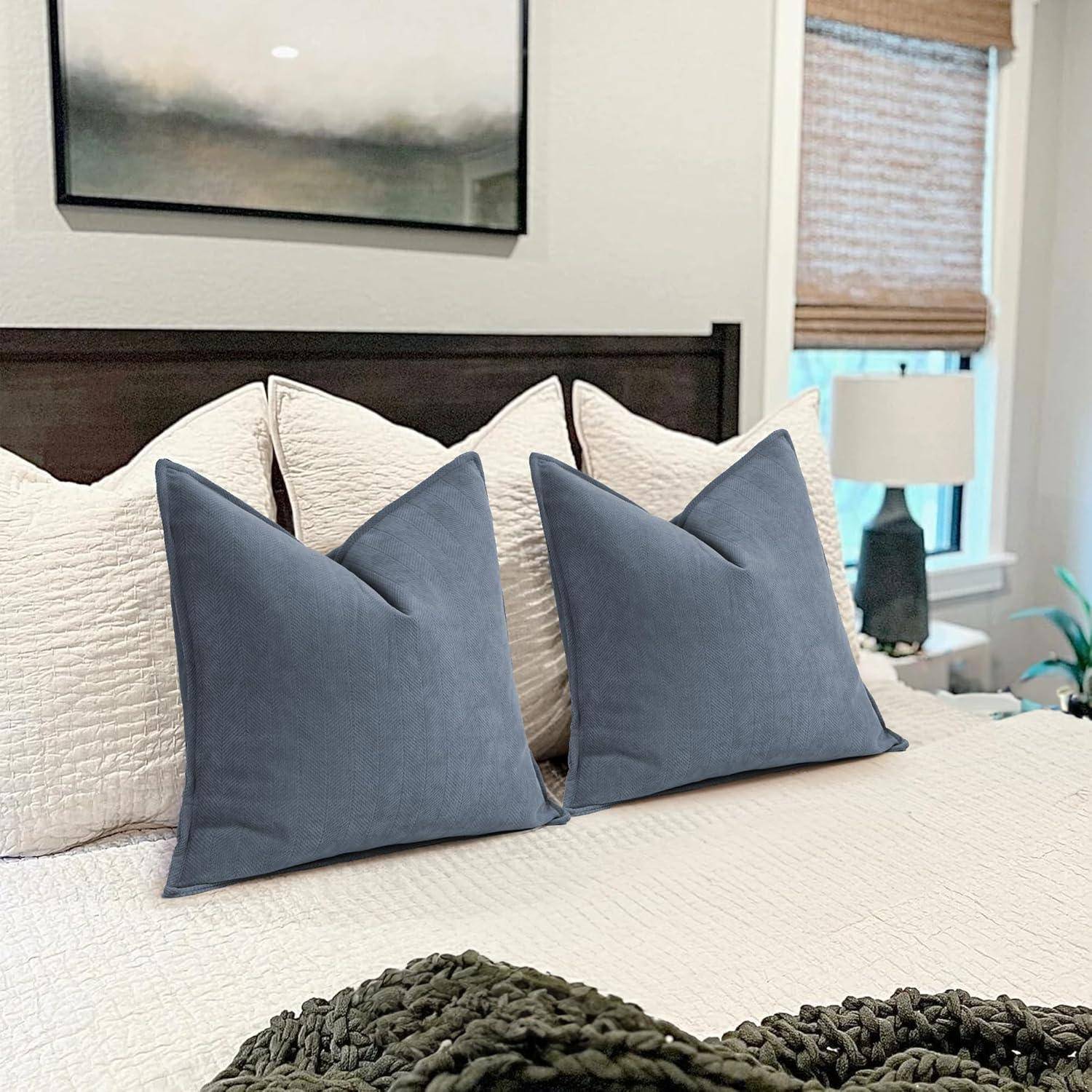Ein ordentlich gemachtes Bett mit vier Kissen – zwei weiße hinten und zwei blaue vorne – unter einer hellgefärbten Decke. Ein Fenster und eine Lampe sind im Hintergrund zu sehen.
