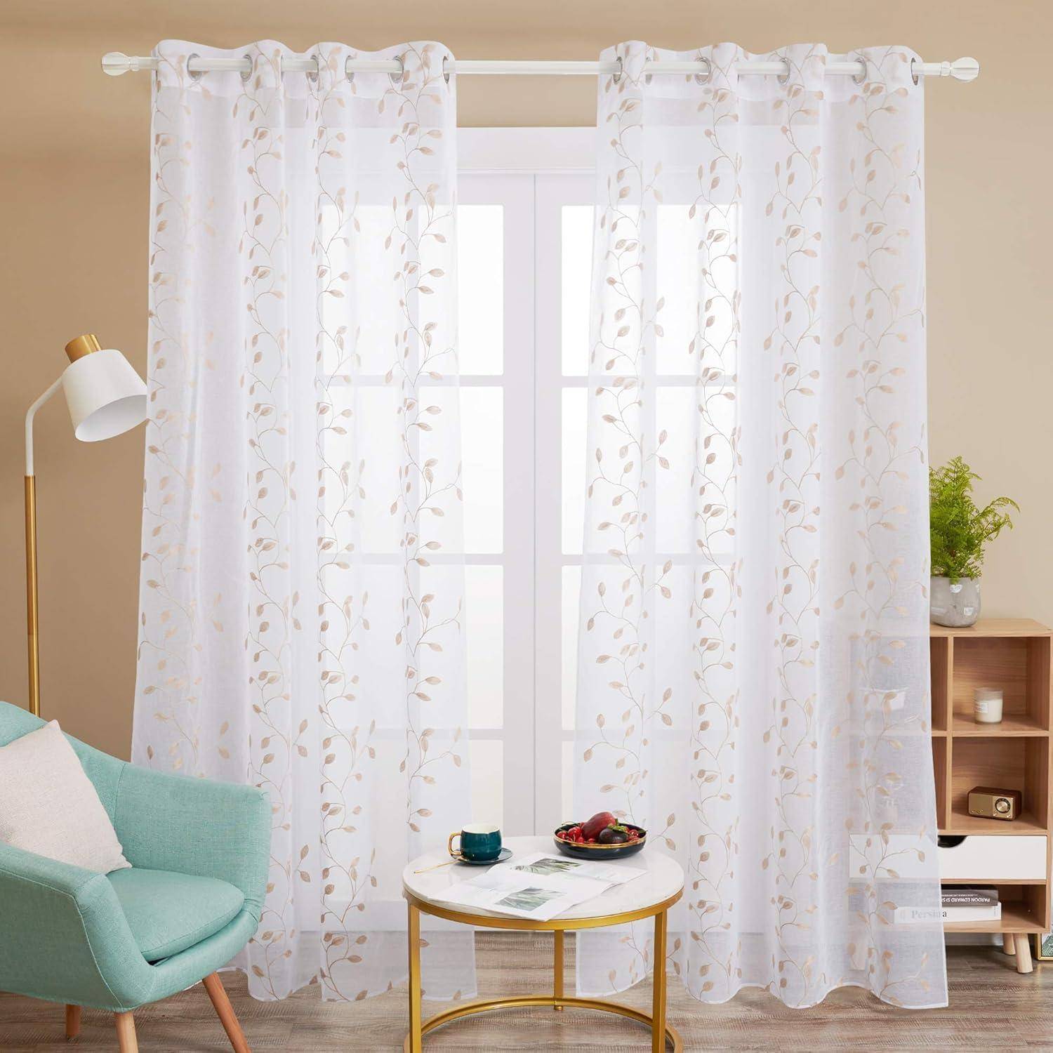Weiße transparente Gardinen mit einem Blattmuster hängen über einem großen Fenster. Ein grüner Stuhl und ein kleiner Holztisch mit zwei Tassen befinden sich in der Nähe.