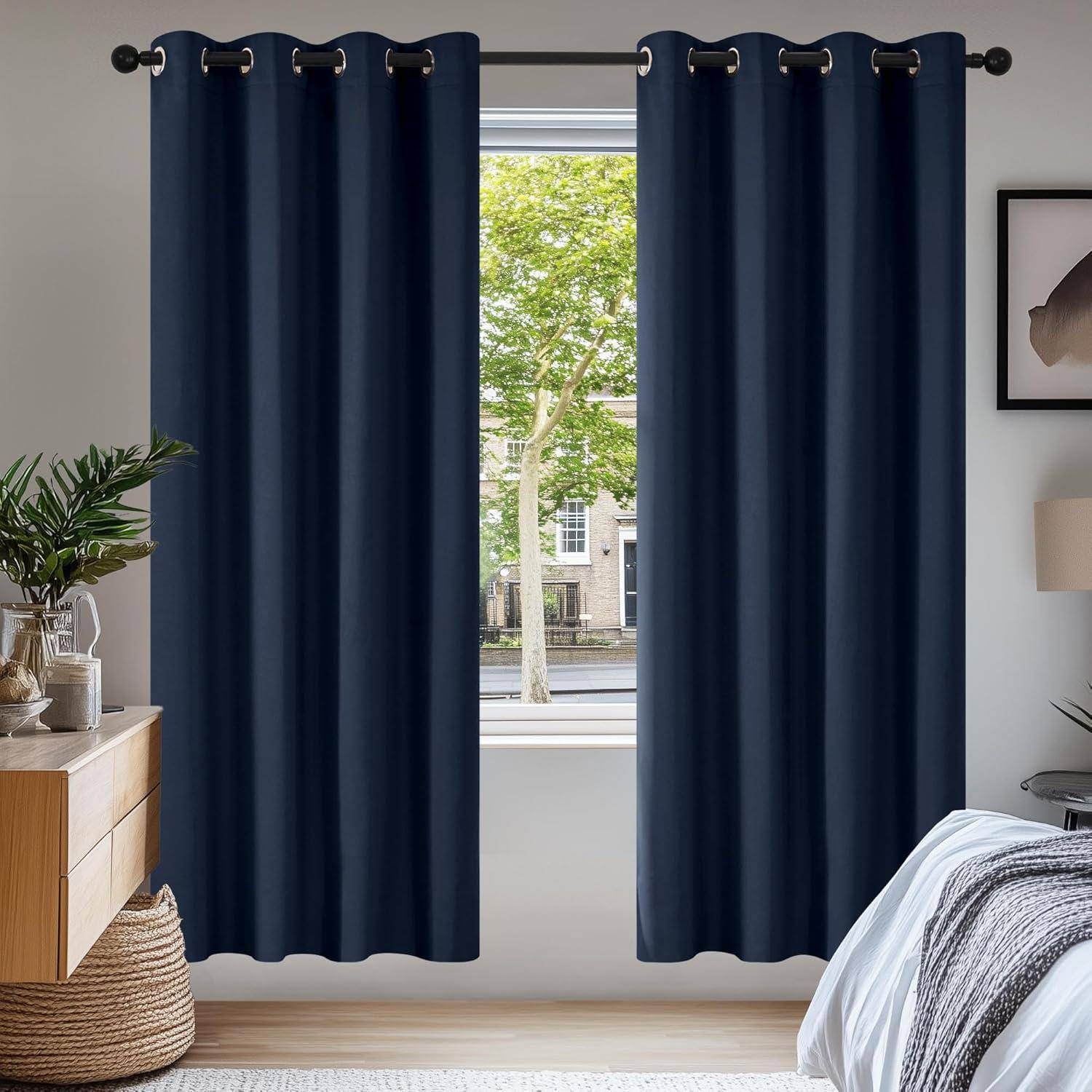 Raum mit dunkelblau Vorhängen, die ein Fenster umrahmen und einen Blick auf Bäume und Gebäude nach draußen freigeben. Ein Holzkommode und ein Bett sind zu sehen.