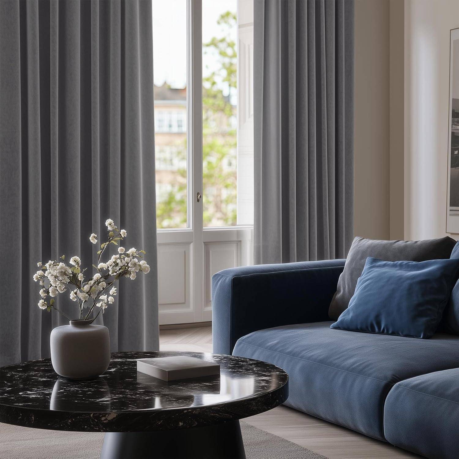Ein gemütliches Wohnzimmer mit einem blauen Sofa und grauen Vorhängen. Ein Marmortisch mit einer Vase mit weißen Blumen steht neben großen Fenstern, die Grünpflanzen zeigen.