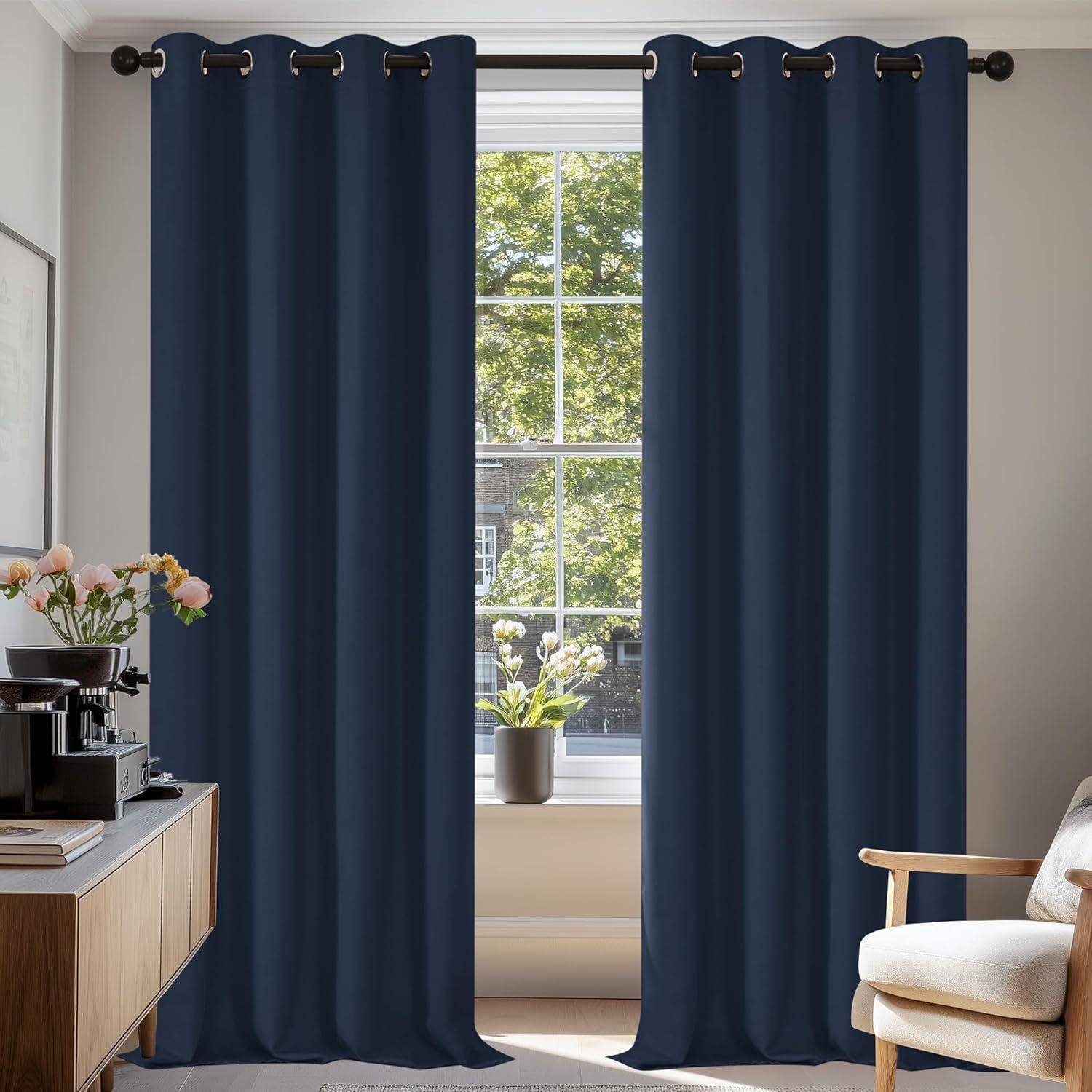 Ein gut beleuchteter Raum zeigt marineblaue Vorhänge, die ein großes Fenster mit Blick auf einen Baum umrahmen. Eine Kaffeeecke und eine Topfpflanze befinden sich in der Nähe.