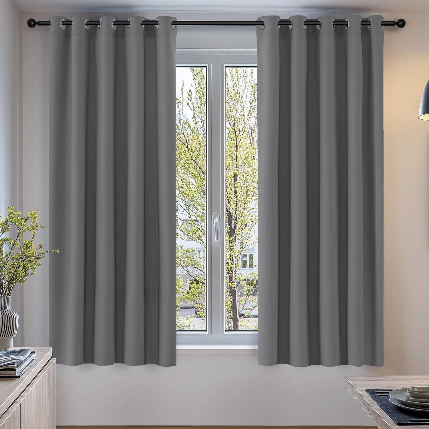 Graue Vorhänge hängen an einer Stange, die ein Fenster rahmen, durch das man Bäume draußen sehen kann. Eine kleine Pflanze und eine Lampe stehen auf einem Tisch neben dem Fenster.