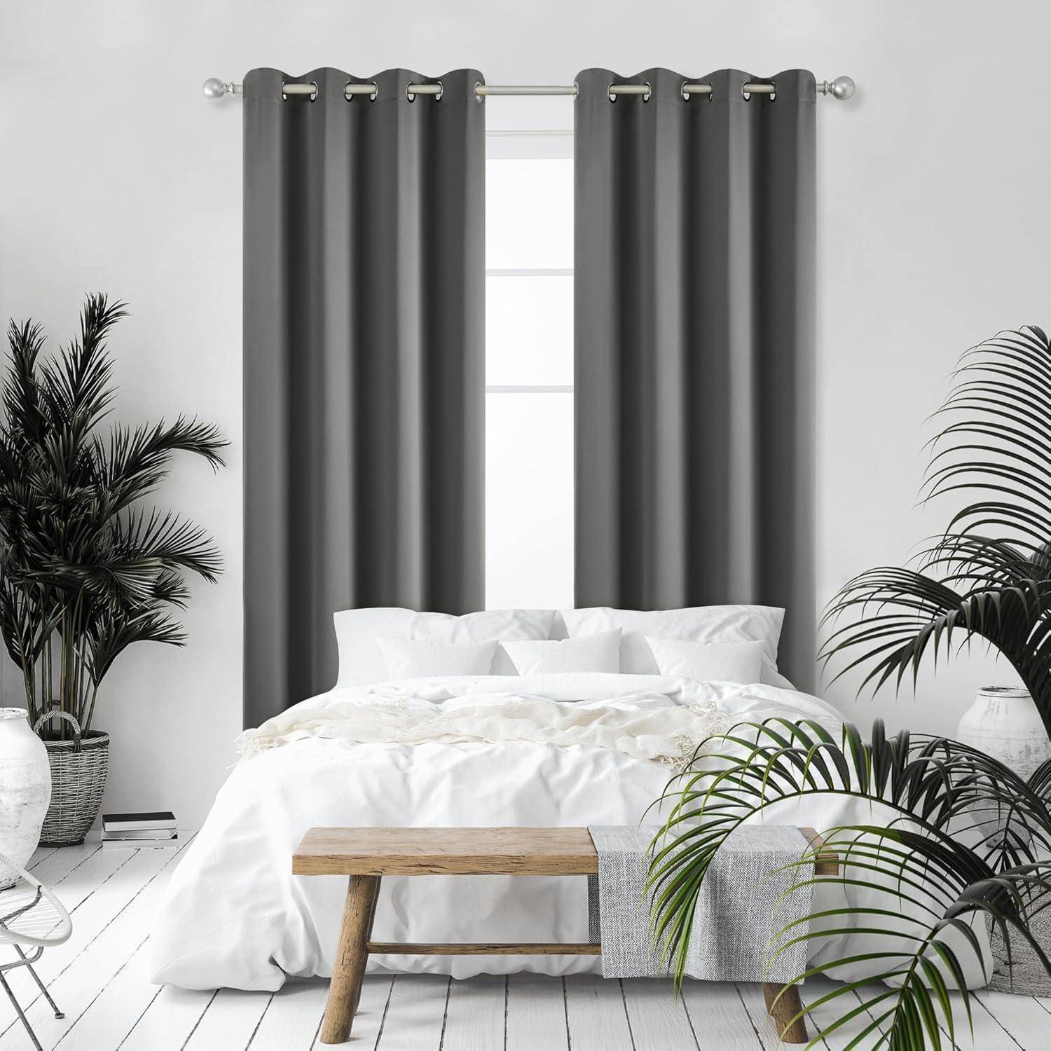 Ein Schlafzimmer zeigt ein ordentlich gemachtes Bett mit weißer Bettwäsche, eine Holzbank am Fußende und zwei graue Vorhänge, die ein Fenster umrahmen, flankiert von Zimmerpflanzen.