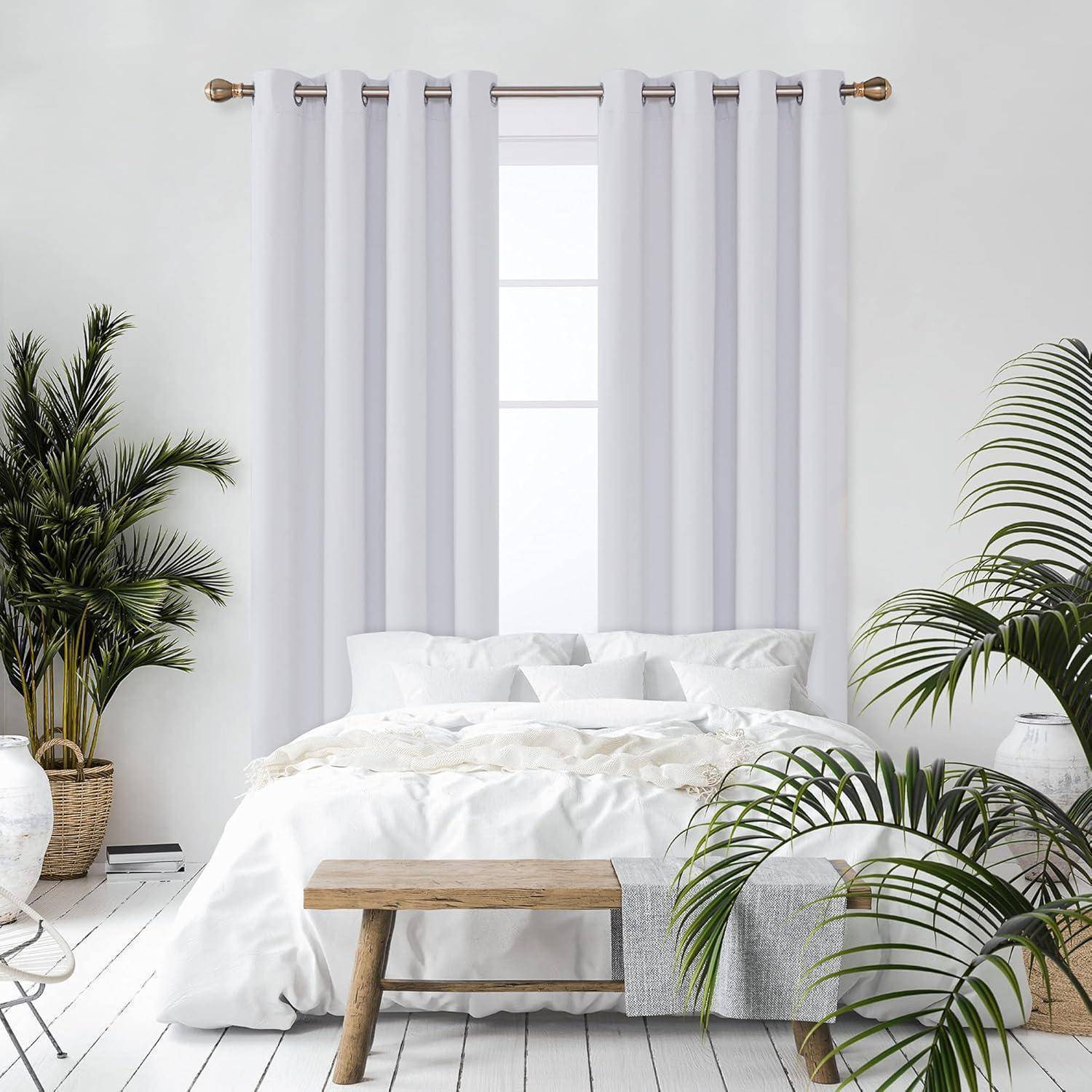 Ein helles Schlafzimmer mit einem weißen Bett, einer hellen Holzbank und einem großen Fenster, das von weißen Vorhängen bedeckt ist. Topfpflanzen befinden sich auf beiden Seiten.