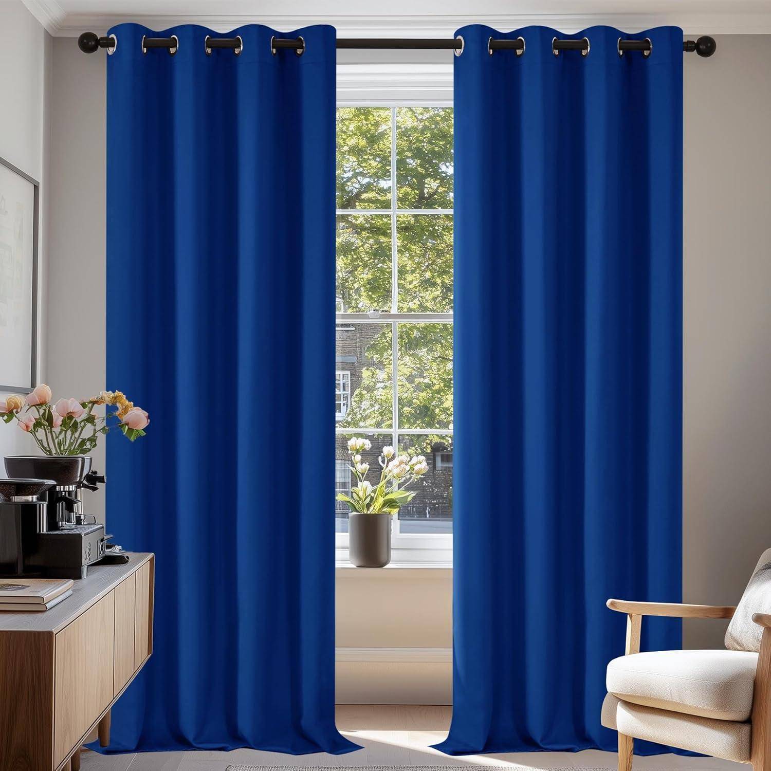 Blaue Vorhänge hängen vor einem großen Fenster mit einer Topfpflanze. Auf der linken Seite steht ein Tisch mit einer Kaffeemaschine und Blumen.