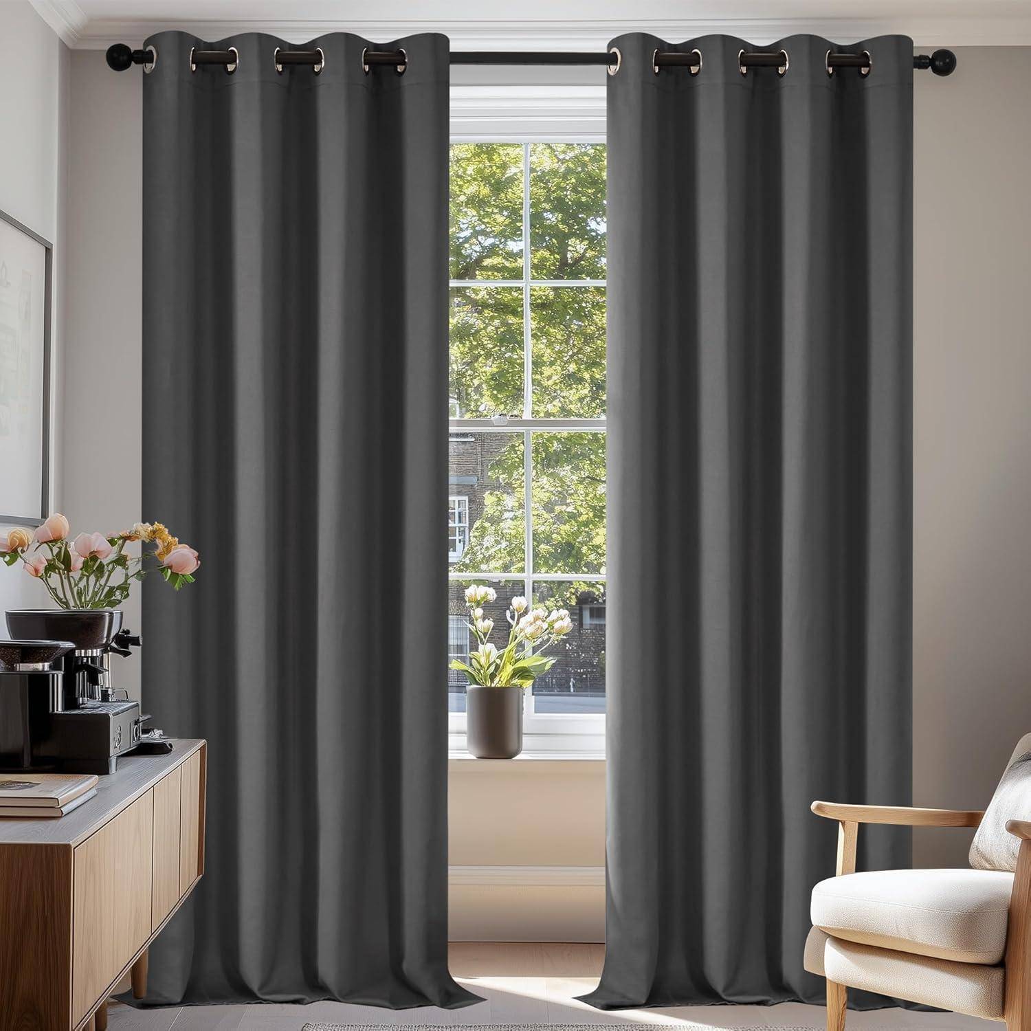 Ein heller Raum mit einem großen Fenster, umrahmt von dunkelgrauen Vorhängen, ein Holzschrank mit Blumen und Büchern links und einem beigen Stuhl rechts.